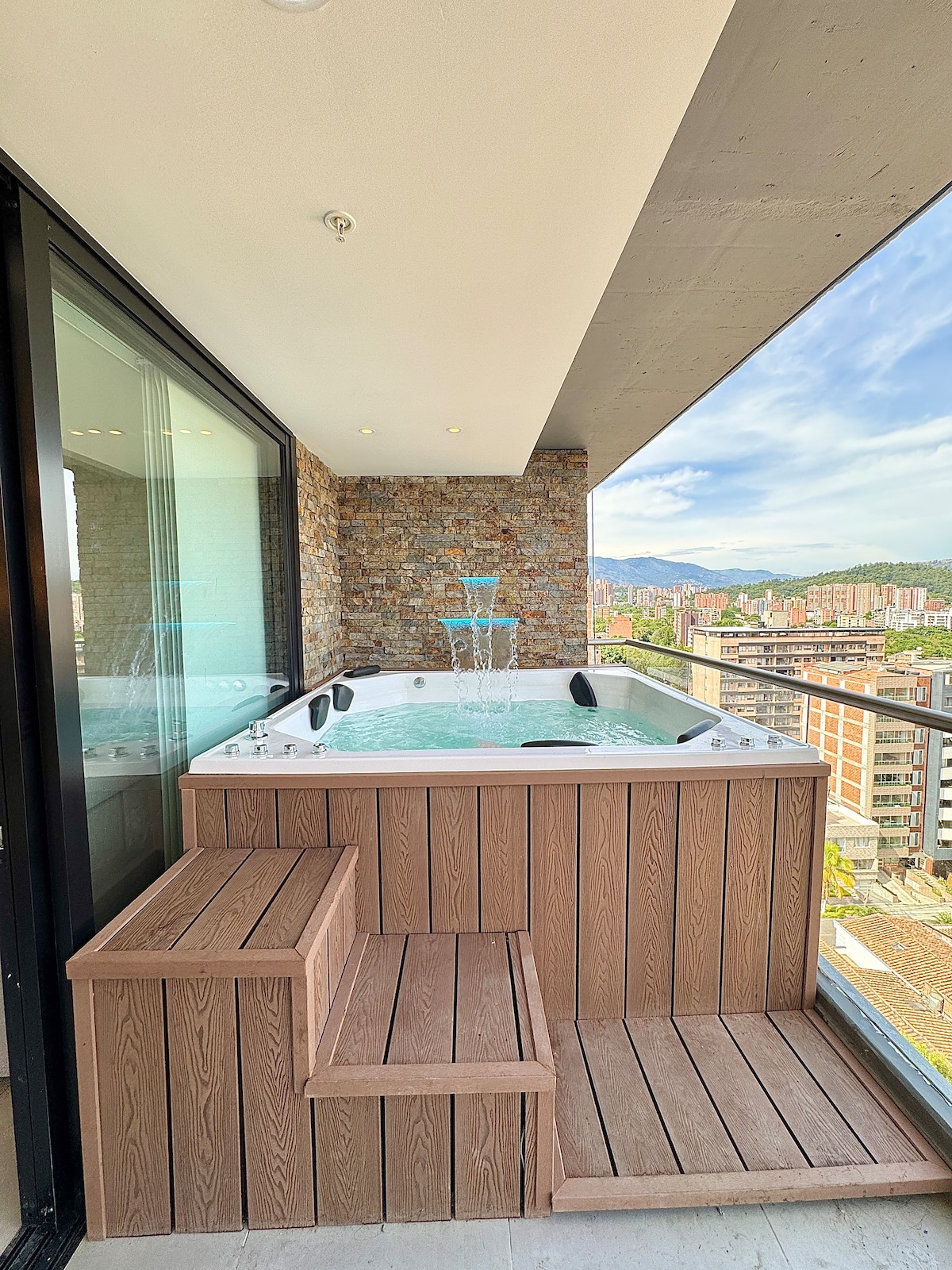A custom jacuzzi is seen on a private balcony, featuring lights and a waterfall feature. Surrounding the jacuzzi are wooden steps leading up to the inviting space. A panoramic view of the city is visible beyond the glass doors, enhancing the outdoor experience.