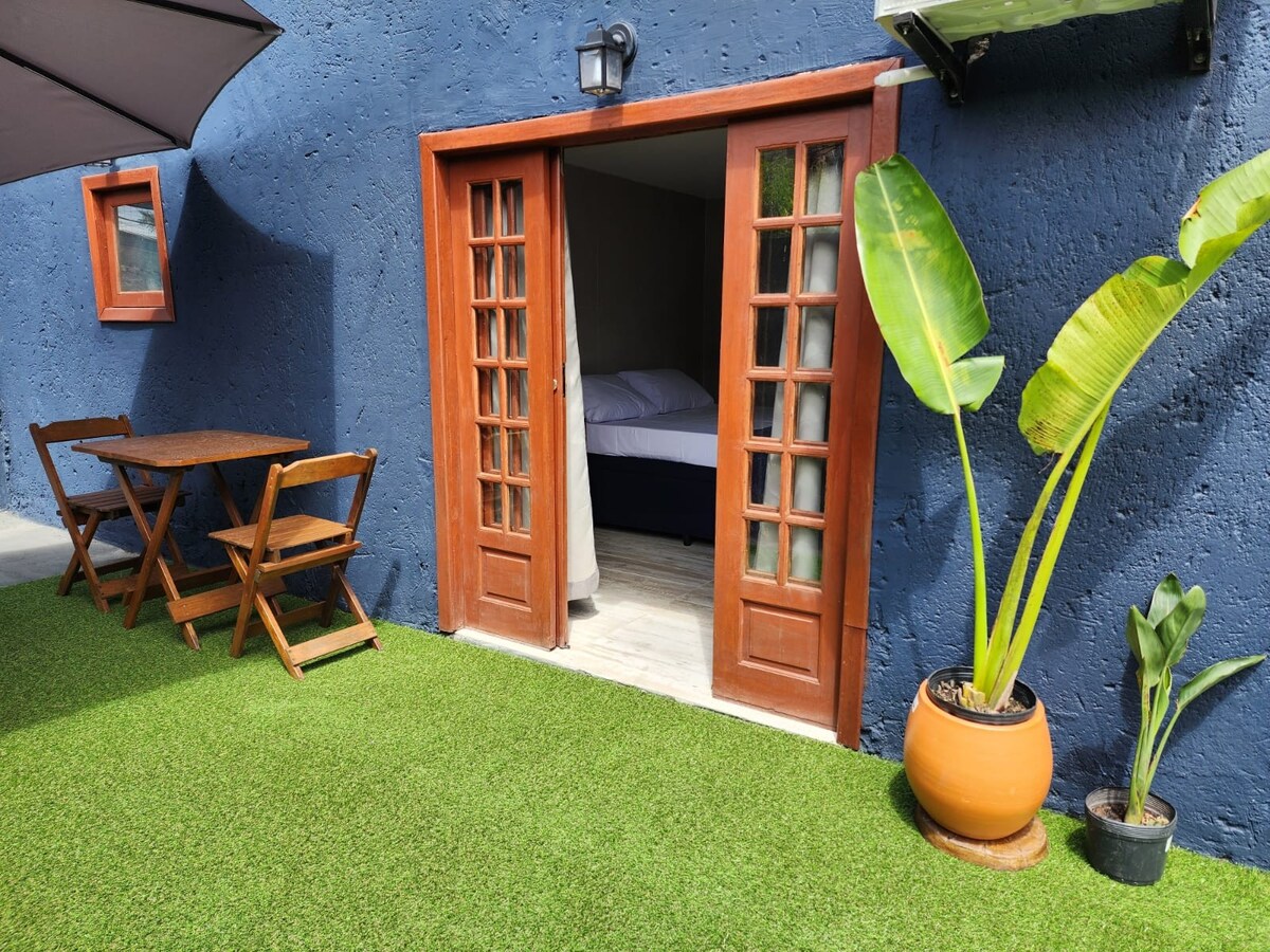 A private outdoor patio features a grassy area with a wooden table and chairs, framed by vibrant green plants. Double doors lead into the cozy interior where a bed is visible. The bold blue walls provide a striking backdrop.