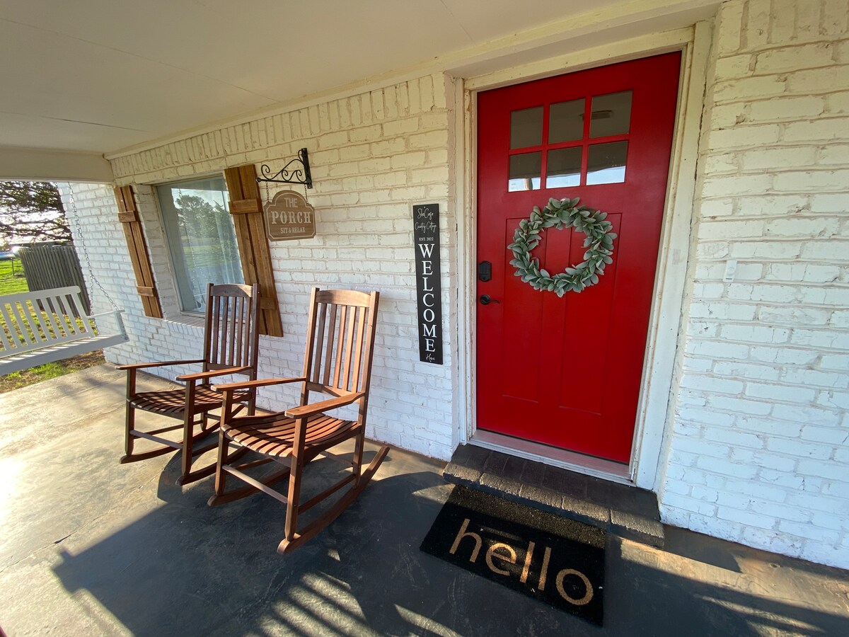 A cheerful red front door is framed by a welcoming wreath and accompanied by a large 'WELCOME' sign. Two wooden rocking chairs sit on the porch beside a doormat featuring the word 'hello'. Natural light spills across the entryway.