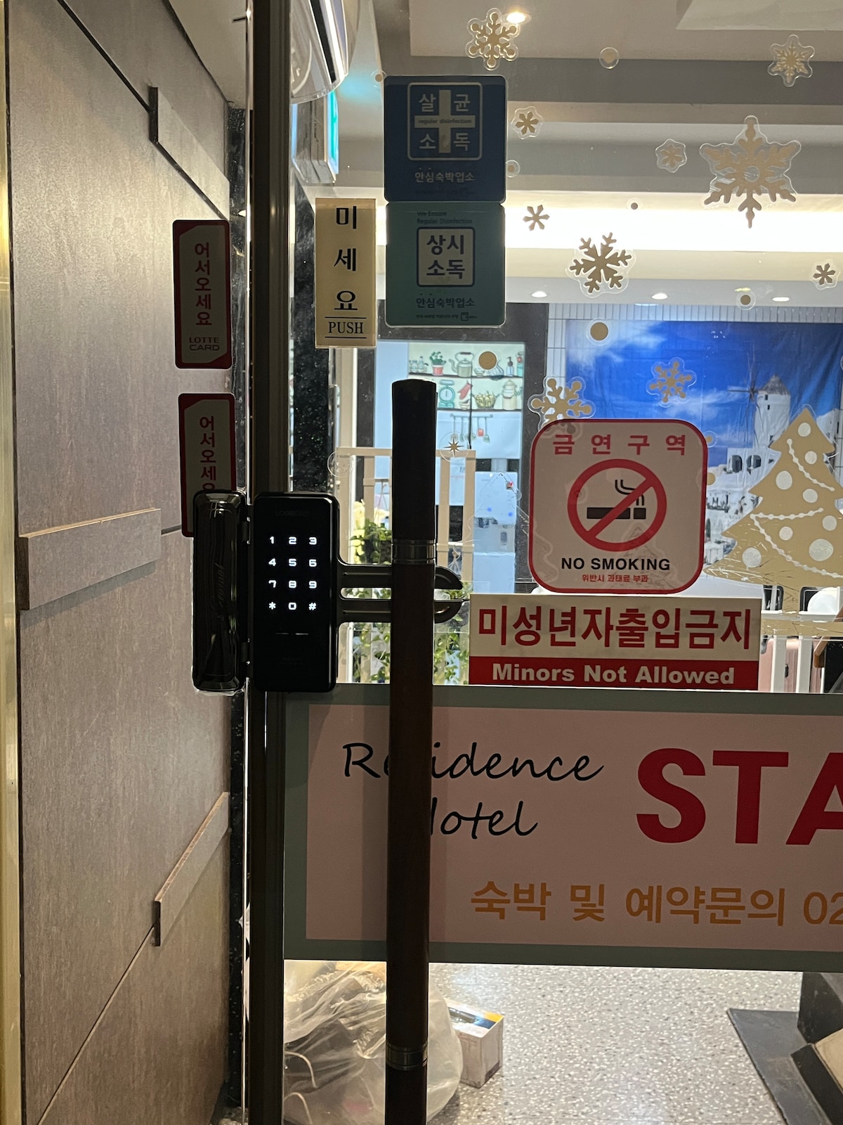 An entryway is depicted, featuring a locked door with a keypad on the right. Various signage is displayed, including instructions for check-in procedures, safety regulations, and a reminder regarding smoking restrictions. Decorative elements are evident, suggesting a welcoming ambiance.