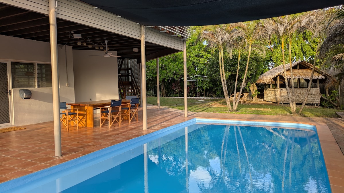 A serene outdoor space features a clear swimming pool reflecting the sky. Adjacent, a dining area with a large table and wooden chairs is positioned under a shaded structure. Lush greenery surrounds the area, with a thatched gazebo visible in the background.