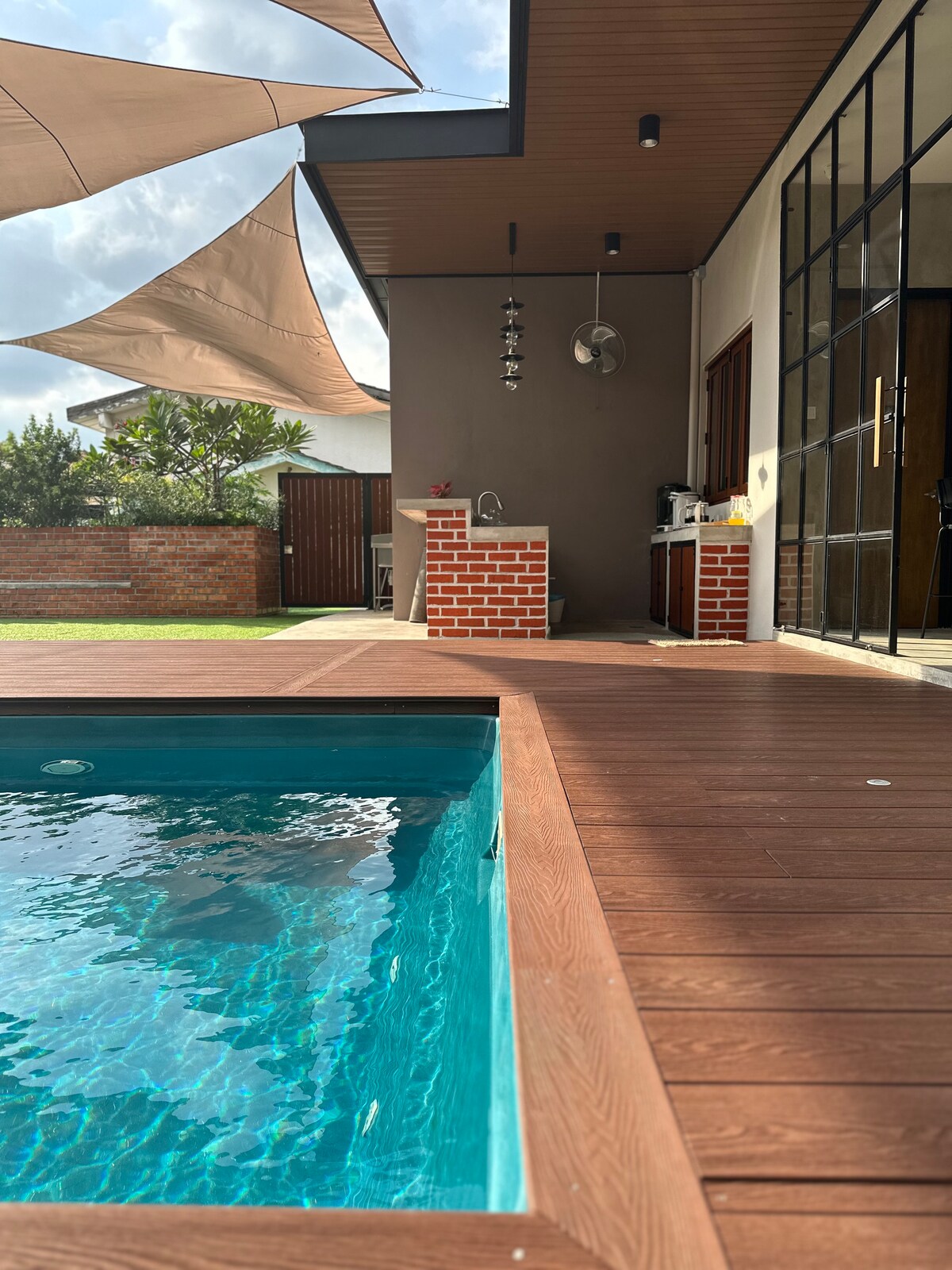 A refreshing shared pool is situated next to a wooden deck, featuring an inviting turquoise hue. An outdoor kitchen area with a brick counter is visible, with a shaded patio above. A well-maintained lawn and a glimpse of the neighboring garden create a serene setting.
