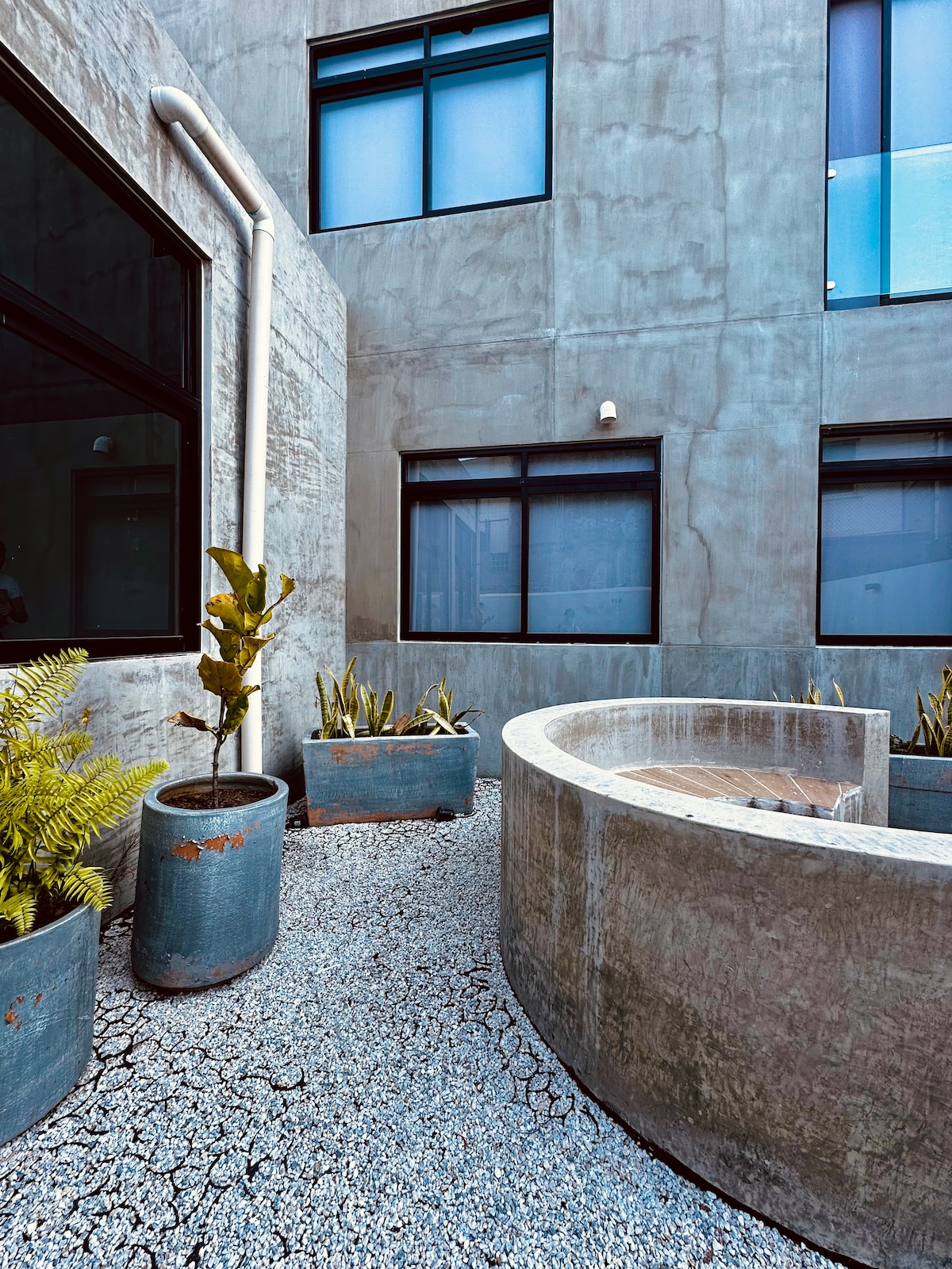 An outdoor space features a circular stone seating area surrounded by minimalist planters with greenery. Light grey walls and large windows create a modern ambiance, while gravel covers the ground for a clean aesthetic.