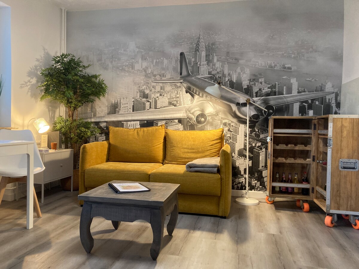 A modern living area features a mustard-yellow sofa, complemented by a wooden table. The wall prominently displays a large black-and-white mural of a vintage airplane over a cityscape. A small plant adds a touch of greenery, while a beverage cart stands nearby.
