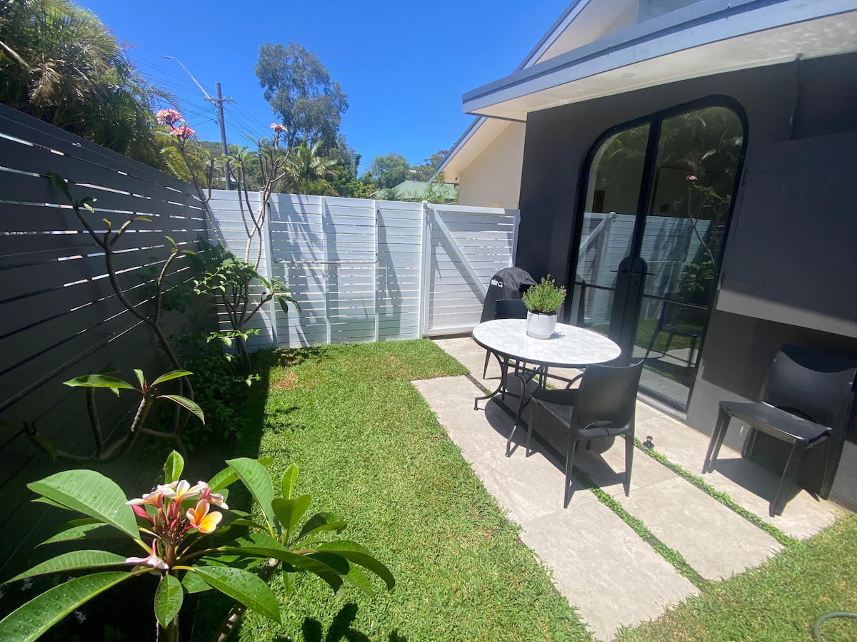 An outdoor seating area is depicted, featuring a round table and two black chairs on a stone patio. Lush green grass surrounds the space, complemented by leafy plants and flowering bushes. A light-colored fence provides privacy while allowing natural light to flow in.
