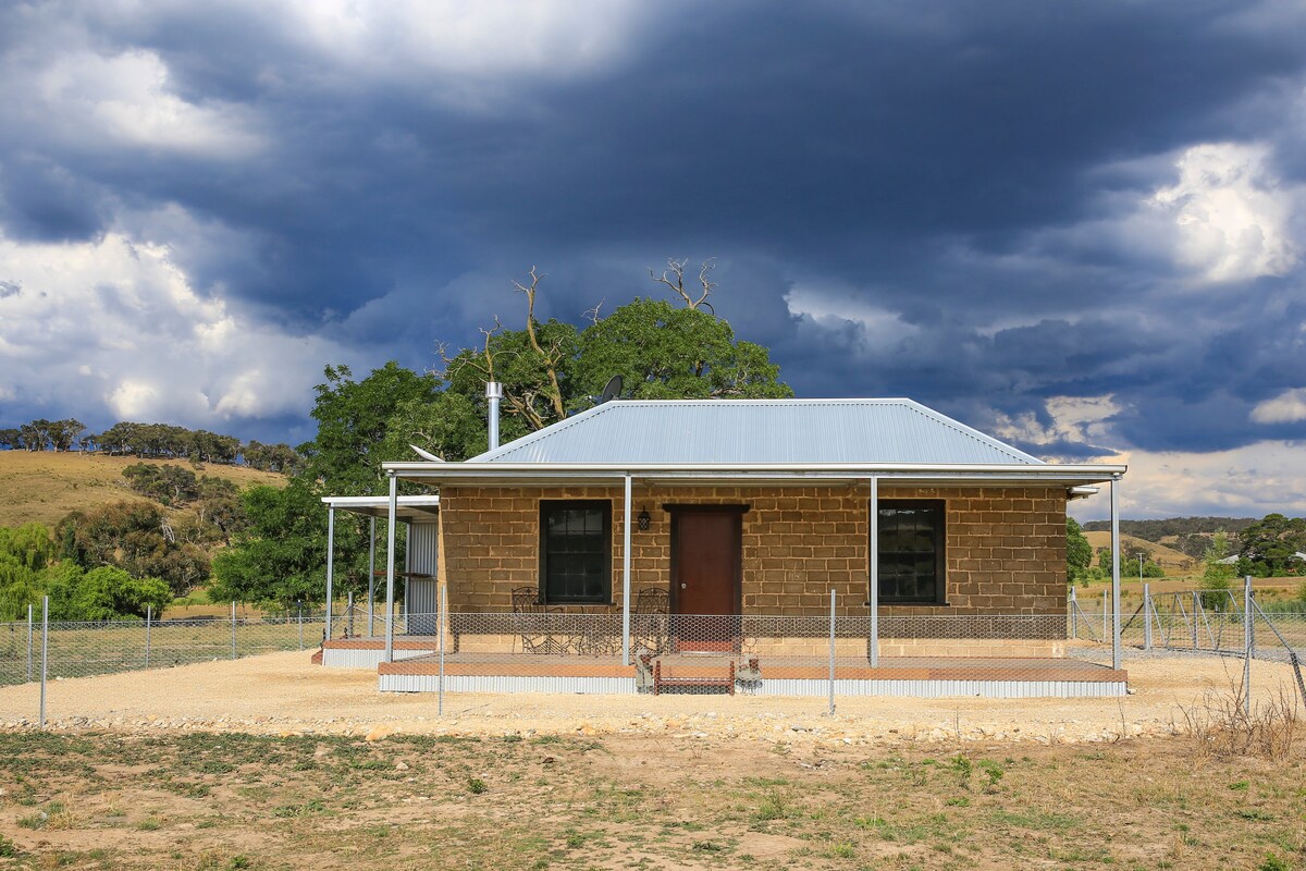 A renovated mud-brick cottage is framed by a rustic fence, set against a dramatic sky with rolling clouds. The structure features a central door, flanked by windows, and a roof with a light blue hue. A peaceful countryside landscape is visible in the background.