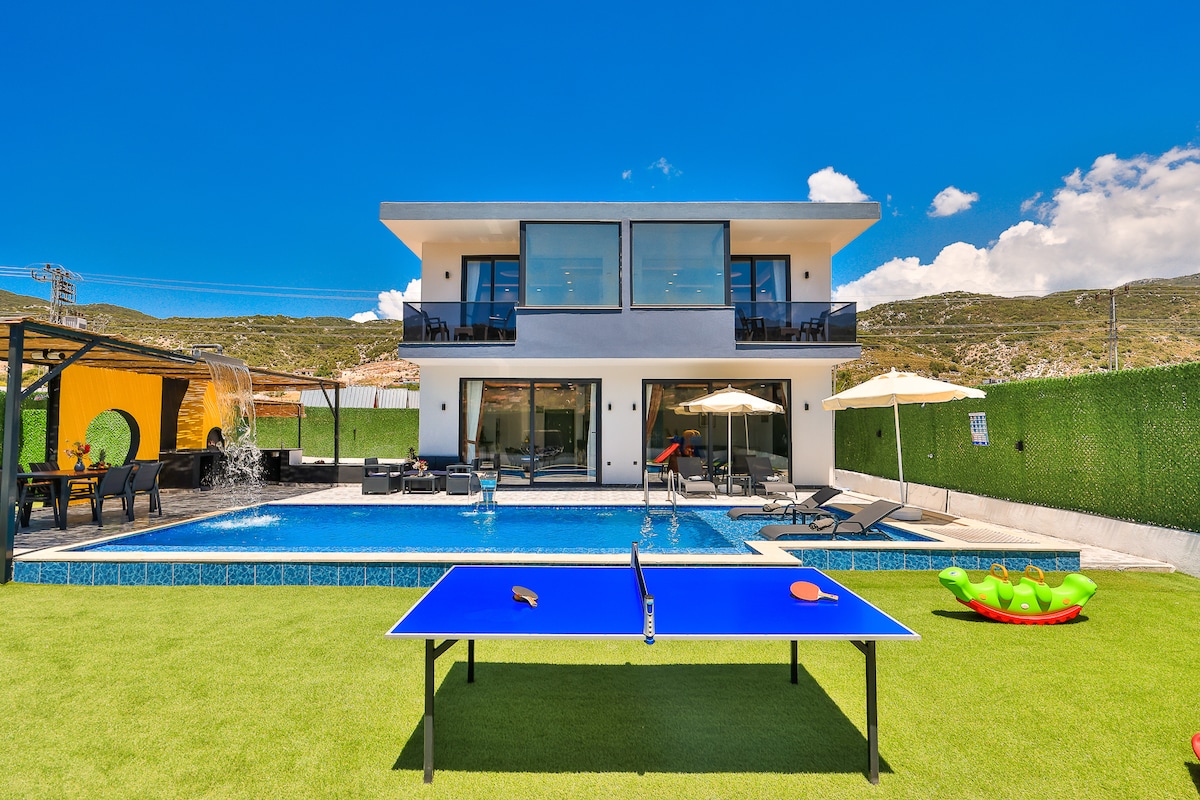 A modern villa is showcased with an inviting pool area, featuring a tennis table positioned on a lush green lawn. Lounge chairs and umbrellas surround the pool, while a covered seating area offers shaded relaxation. The facade displays large glass doors, enhancing natural light.