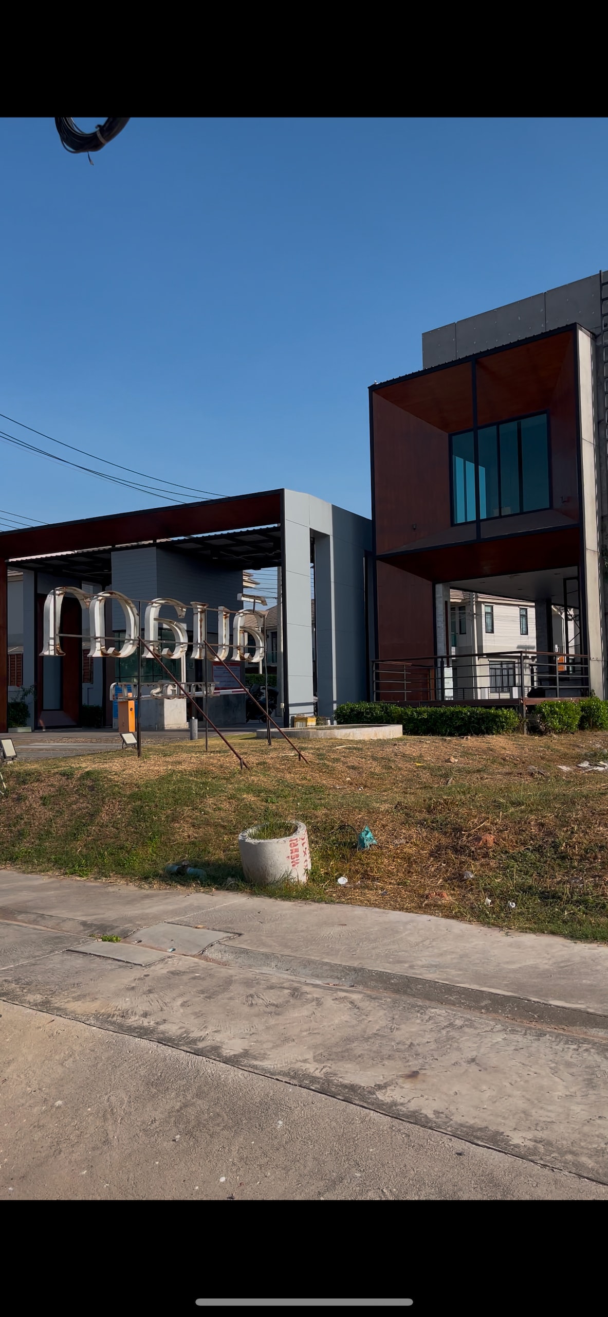 A modern building structure with a combination of sleek lines and large windows is showcased. The facade includes both metal and glass elements, while a sign is clearly visible at the entrance. The area surrounding the property features grass and paved pathways.