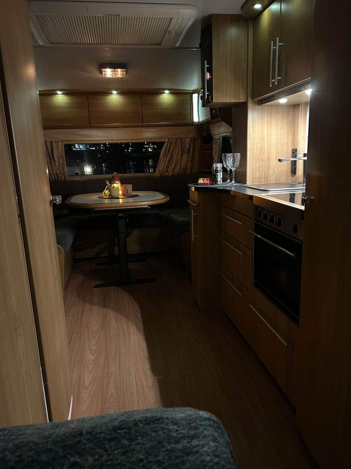 A compact interior of a campervan is visible, featuring wood paneling and soft lighting. A small dining table with two seats is positioned beside a window, while a kitchenette with a stove and cabinets is situated across from it. The atmosphere is calm and cozy.