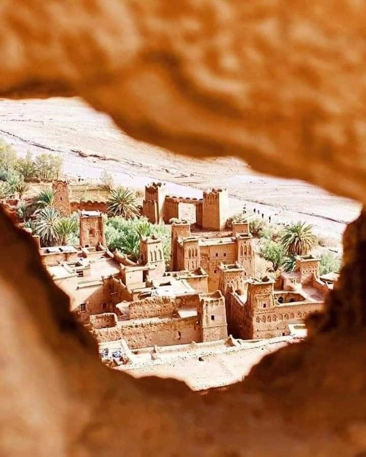 An aerial view of a traditional village reveals a maze of earthen structures interspersed with lush palm trees. The warm tones of the buildings contrast with the surrounding natural landscape, showcasing the rich texture of local architecture framed by a natural opening.