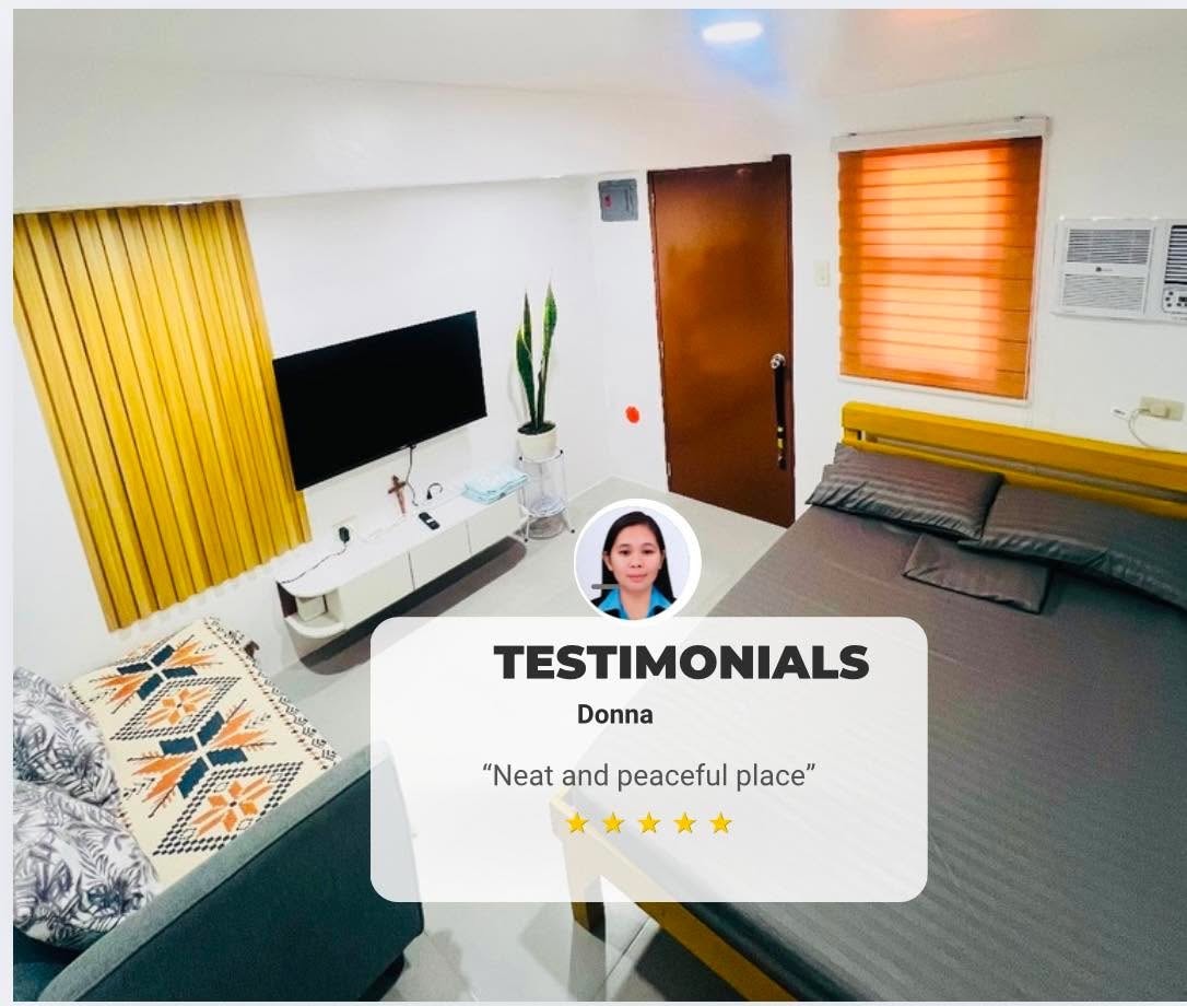 A tidy studio space is displayed with a compact sleeping area featuring a spacious bed covered in gray bedding. A flat-screen TV is mounted on the wall opposite, with modern decorations including a potted plant and a patterned throw pillow on a chair.