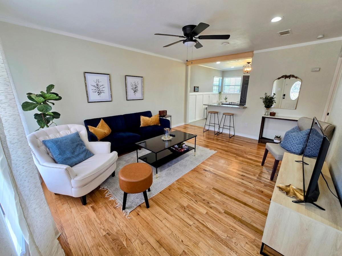 A comfortable living area features a cozy white armchair, a navy blue sofa, and a round ottoman. A glass coffee table sits atop light hardwood flooring, surrounded by framed wall art. A ceiling fan and natural light from windows enhance the inviting space.