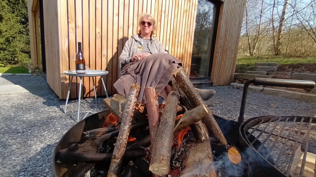 A fire pit is surrounded by neatly arranged logs, with flames visible among the wood. A small side table stands next to it, positioned beside a rustic wooden structure. Sunlight filters through trees in the background, illuminating the gravel area.