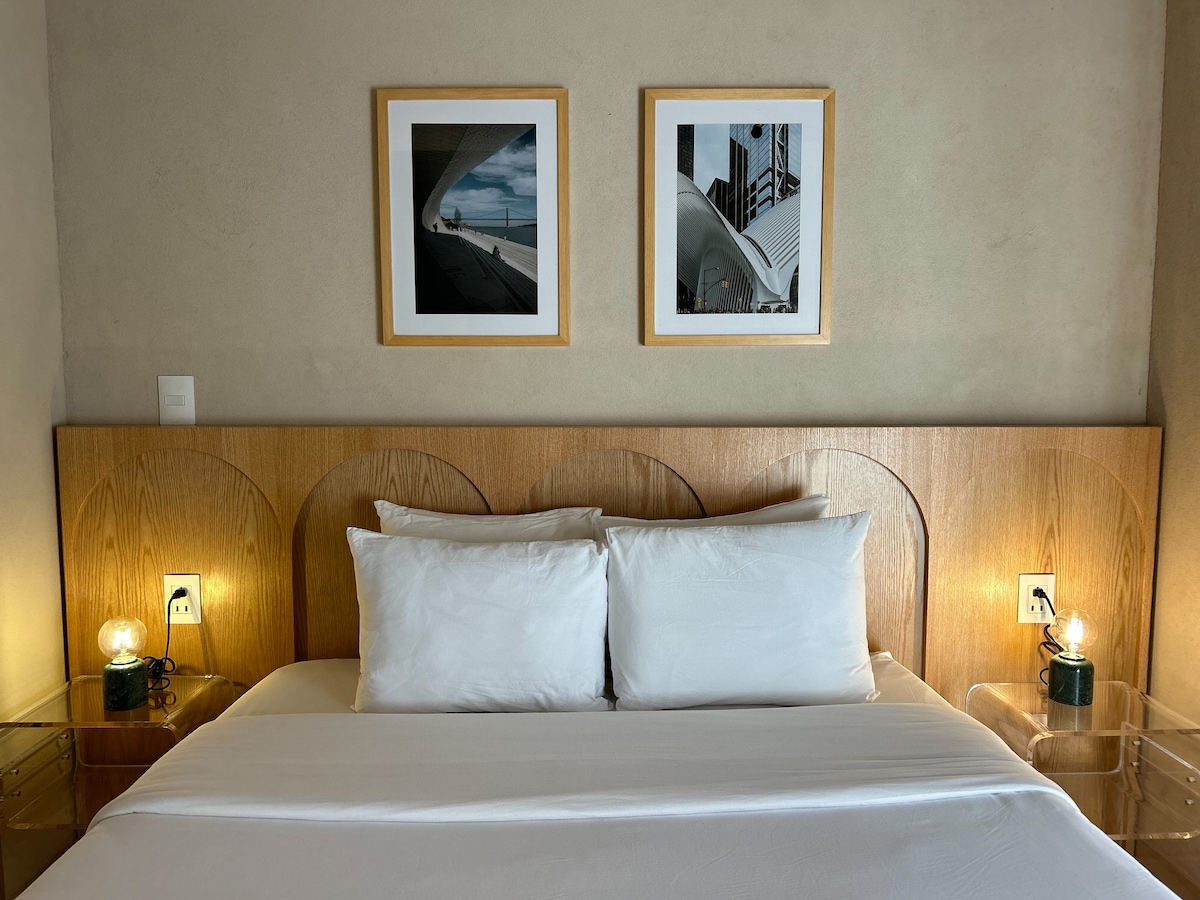 A cozy sleeping area features a spacious bed with white linens and two decorative pillows. Above the bed, two framed photos are displayed on the wall. Nightstands with gentle lighting flank the bed, creating a serene and inviting environment.