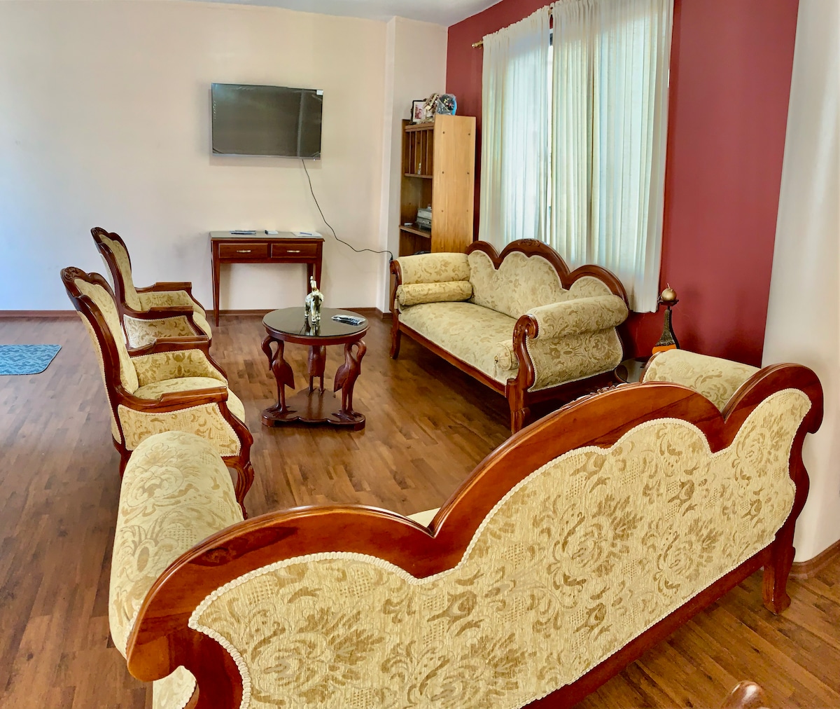 A spacious living area features a traditional seating arrangement with cream-colored upholstered sofas and matching armchairs. A wooden coffee table sits at the center, complemented by a decorative vase. Natural light filters through large windows, enhancing the warm tones of the wooden flooring.