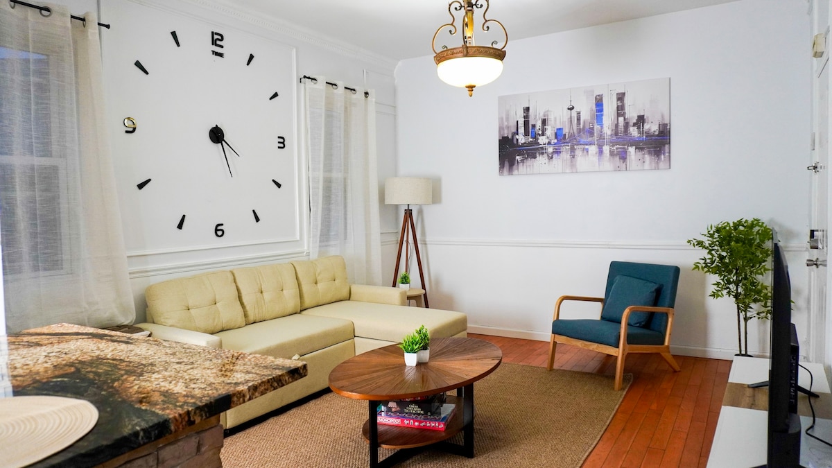 A spacious living room is highlighted by a cozy sofa facing a coffee table. A large wall clock is displayed prominently above the sofa. Nearby, a blue accent chair complements the modern aesthetic. Natural light filters through the sheer curtains, creating a bright atmosphere.