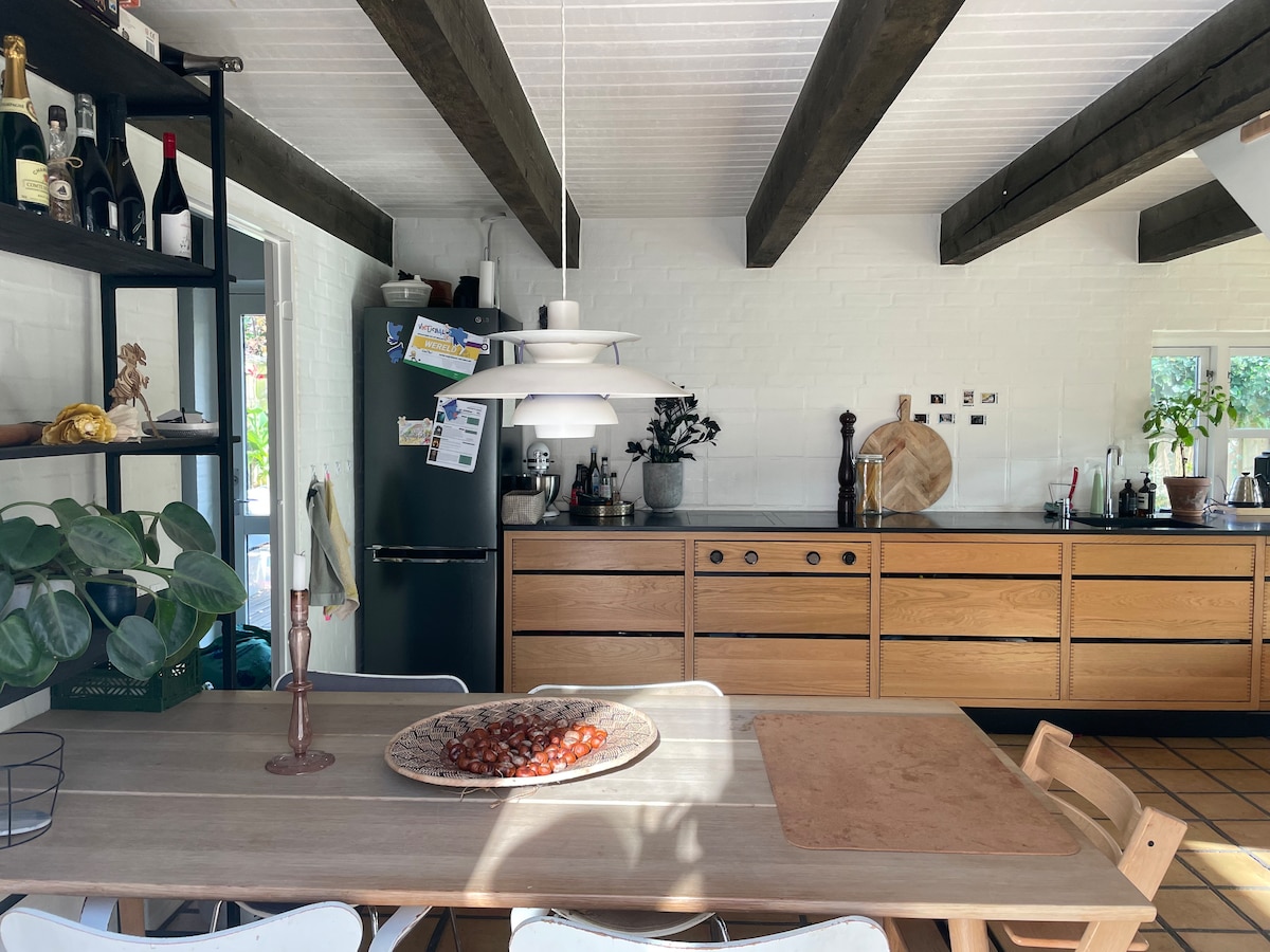 A modern kitchen with wooden cabinetry is highlighted by natural light streaming through windows. A dining table, set with a decorative bowl, is accompanied by what appears to be a mix of wooden and metal chairs. Dark beams contrast with the white walls, creating a welcoming space.