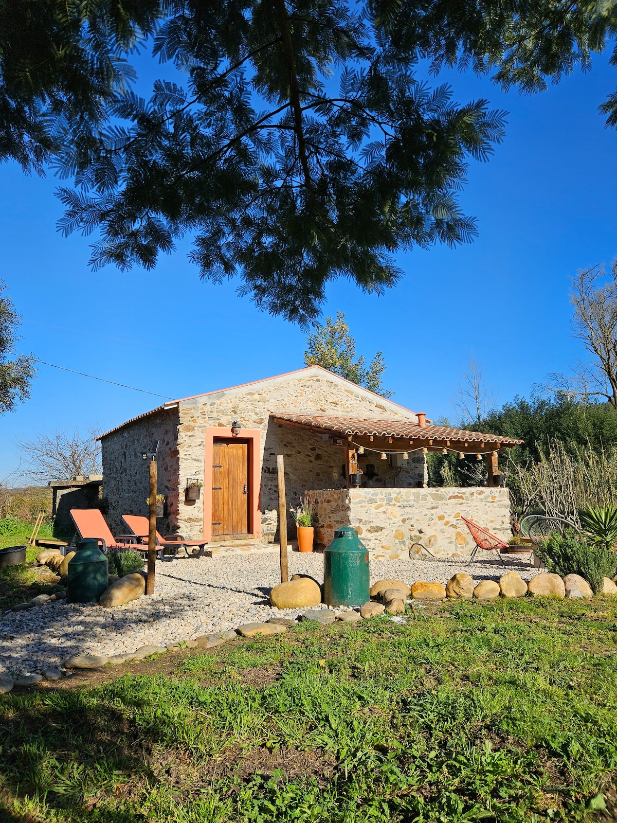 A charming stone watermill is nestled in a lush green landscape, featuring a rustic exterior with wooden doors and a tiled roof. Outdoor seating is provided by two lounge chairs and a sitting area, surrounded by natural stones and vibrant vegetation.