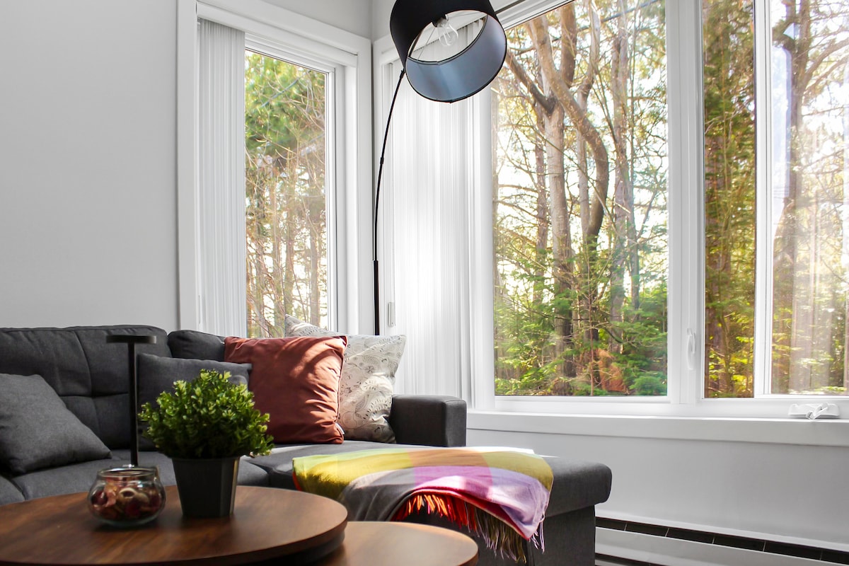 A comfortable seating area is highlighted by a dark grey sofa adorned with plush cushions and a vibrant throw blanket. Large windows offer a view of surrounding trees, allowing natural light to brighten the space. A round wooden table with a decorative plant and glass container adds a touch of warmth.