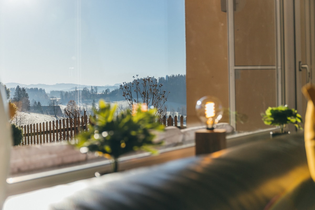 A sunlit view from the living area reveals the distant mountains and a serene landscape. The image includes a bright light bulb on a wooden base and decorative green plants positioned near the window, creating a sense of openness and connection with nature.