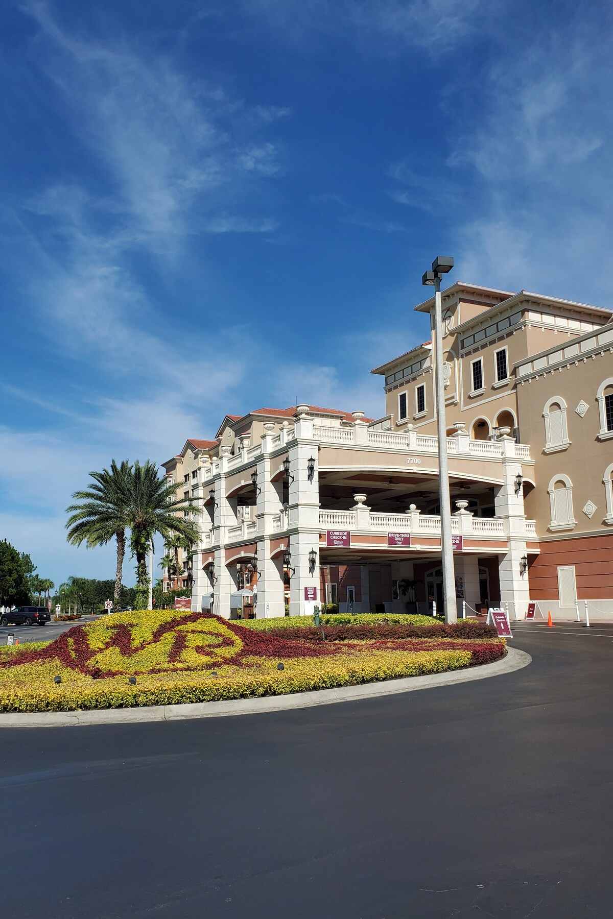 The entrance of the resort is framed by vibrant landscaping featuring colorful flowers arranged to form the initials 'VB.' A multi-story structure with decorative architecture and palm trees is visible against a clear blue sky.