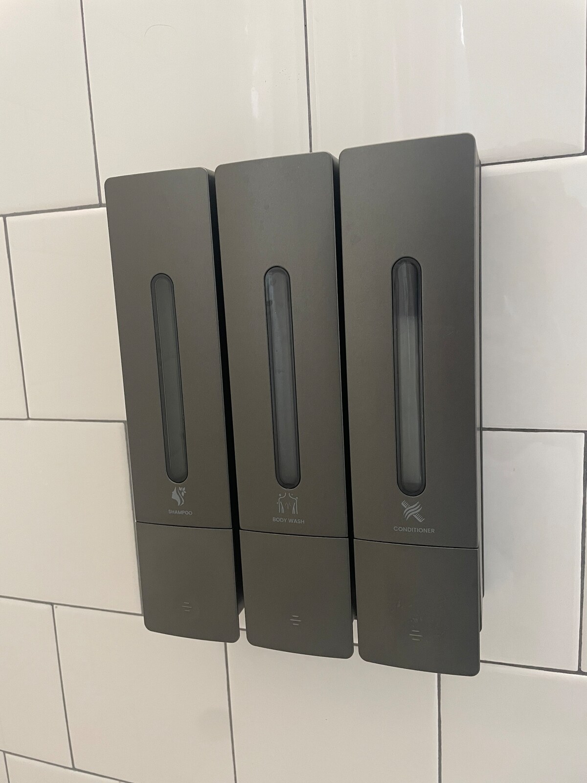 A modern wall-mounted dispenser is shown, containing three compartments labeled for different amenities. The sleek design and minimalistic gray finish blend seamlessly with the surrounding white tiled wall.