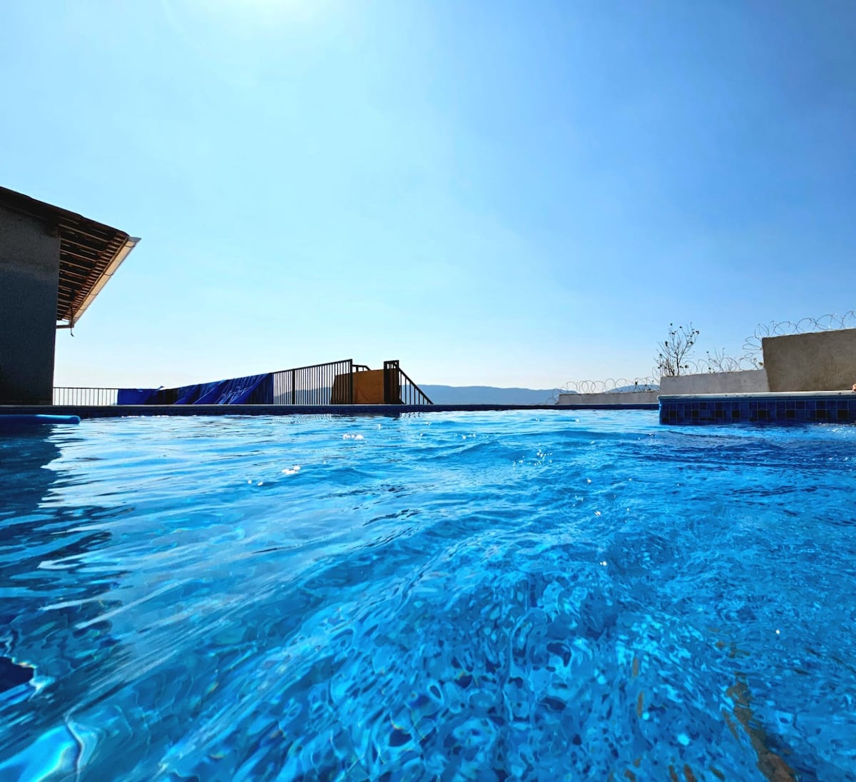 A clear blue swimming pool reflects sunlight, surrounded by a serene rooftop area. The horizon shows distant mountains under a cloudless sky, enhance tranquility. The pool's water appears inviting, with gentle ripples across the surface.