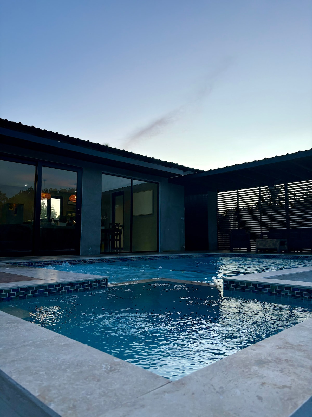 A serene outdoor area features a sparkling pool surrounded by sleek stone tiles. Large glass doors offer views into the interior space, while a partially shaded terrace provides additional seating options. The evening sky casts a soft glow over the tranquil setting.
