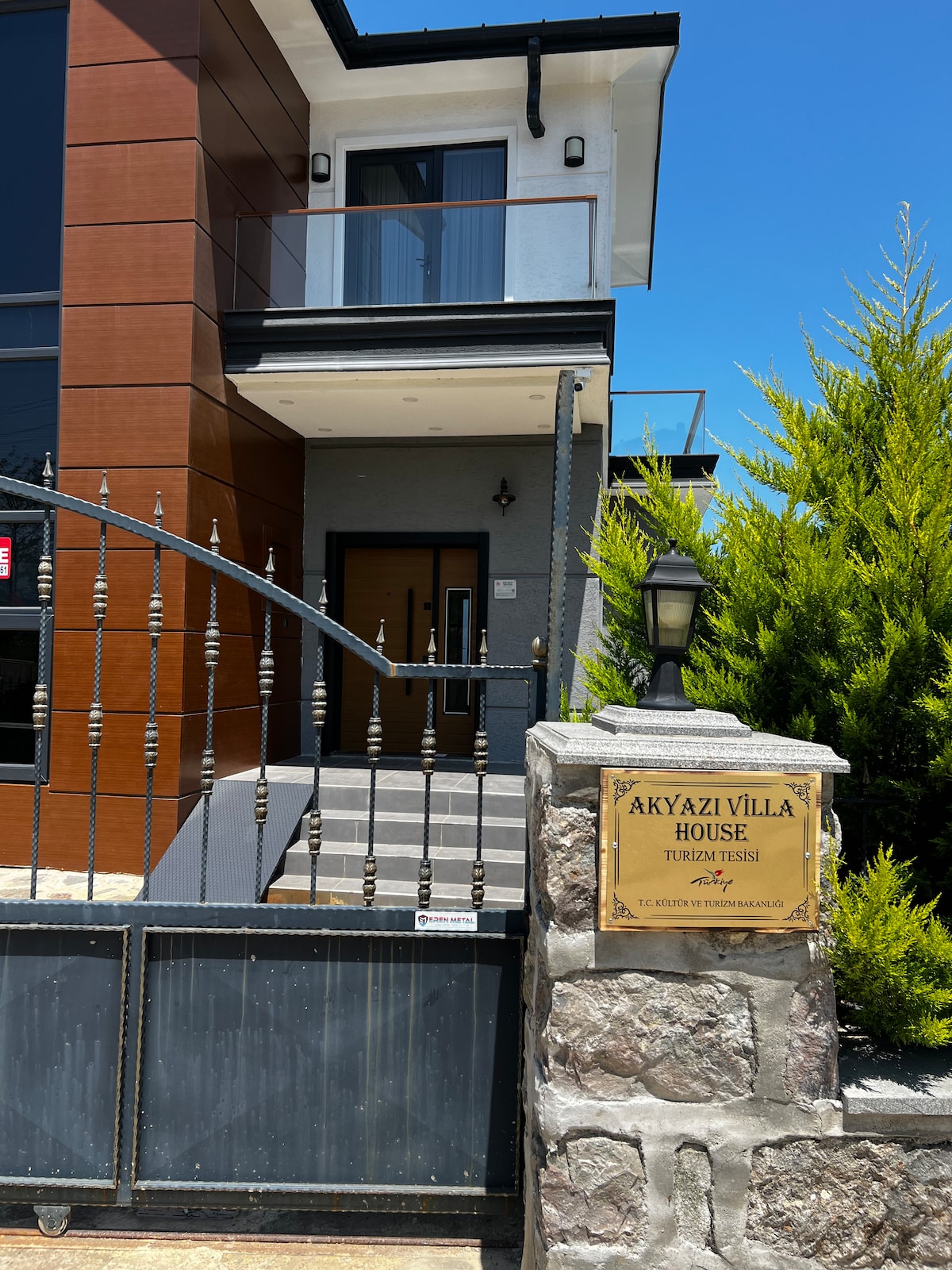 The entrance of Akyazı Villa features a combination of smooth stone and wooden surfaces, with a gated entryway. A sign displaying the villa's name is prominently displayed on a stone pillar beside the gate, surrounded by well-maintained greenery.