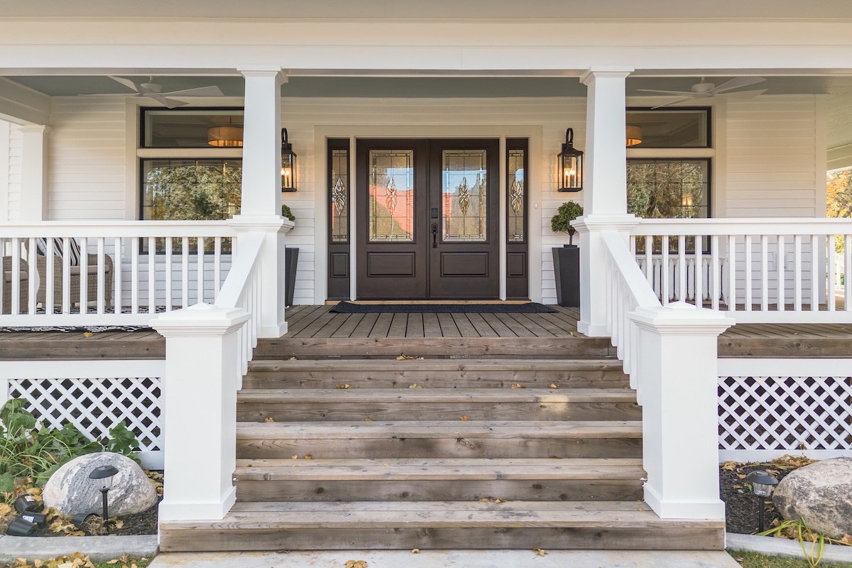 The inviting entrance features a set of double doors framed by large lanterns. Wooden steps lead up to a broad porch, adorned with white railings. Flanking the entrance are potted plants, providing a touch of greenery amidst the wooden and white façade.