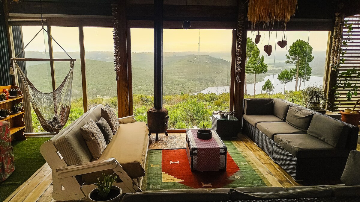 A cozy living area is furnished with a mix of seating options, including a hammock and sectional sofas. Large windows offer expansive views of the surrounding landscape, with rolling hills and trees visible in the distance. A small stove adds to the cabin's warmth.