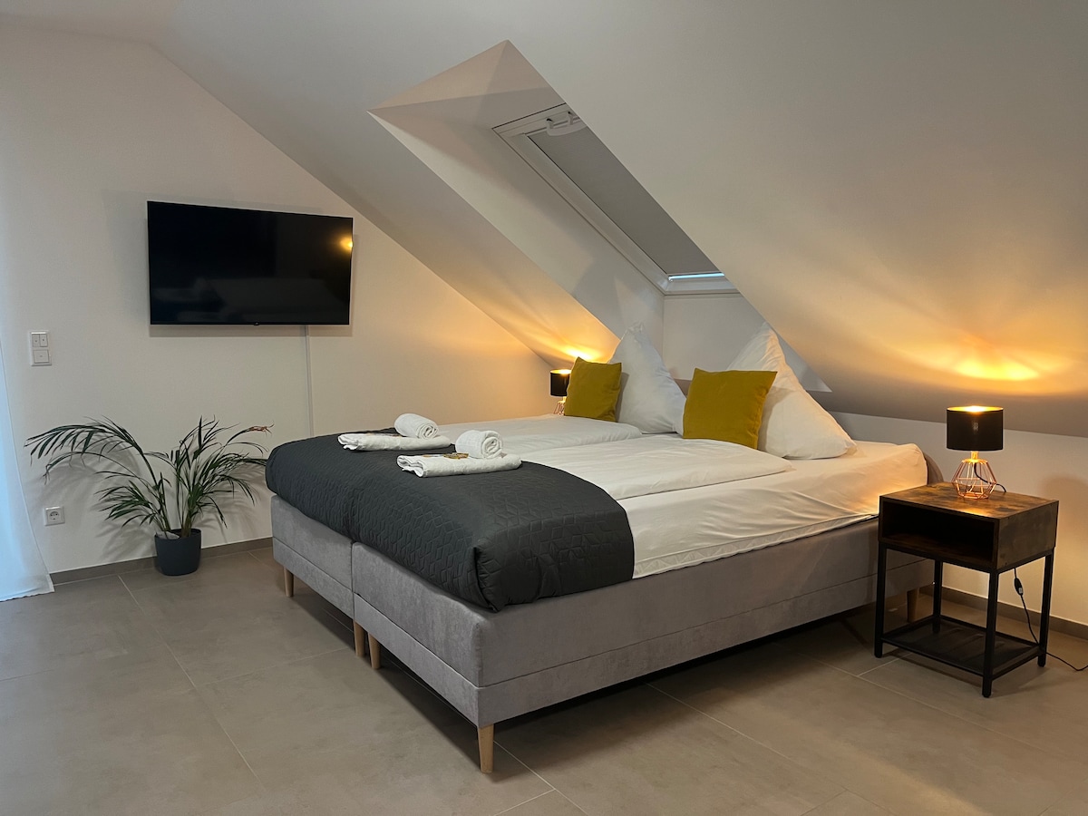 A cozy sleeping area features a spacious bed adorned with soft pillows, complemented by warm ambient lighting from bedside lamps. A flat-screen TV is mounted on the wall, and a potted plant adds a touch of greenery to the corner.