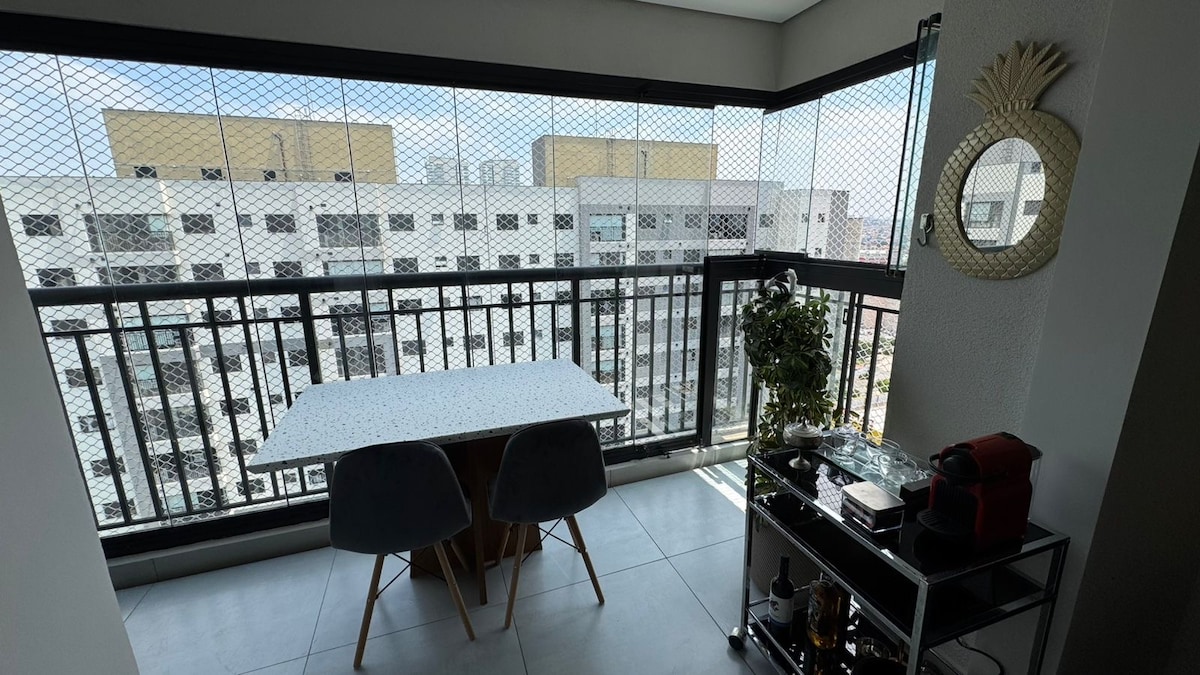A balcony space features a table with four chairs, complemented by a decorative plant. The metal and glass construction provides a clear view of the exterior, with a protective netting in place. A small cart holds a coffee machine and additional items.