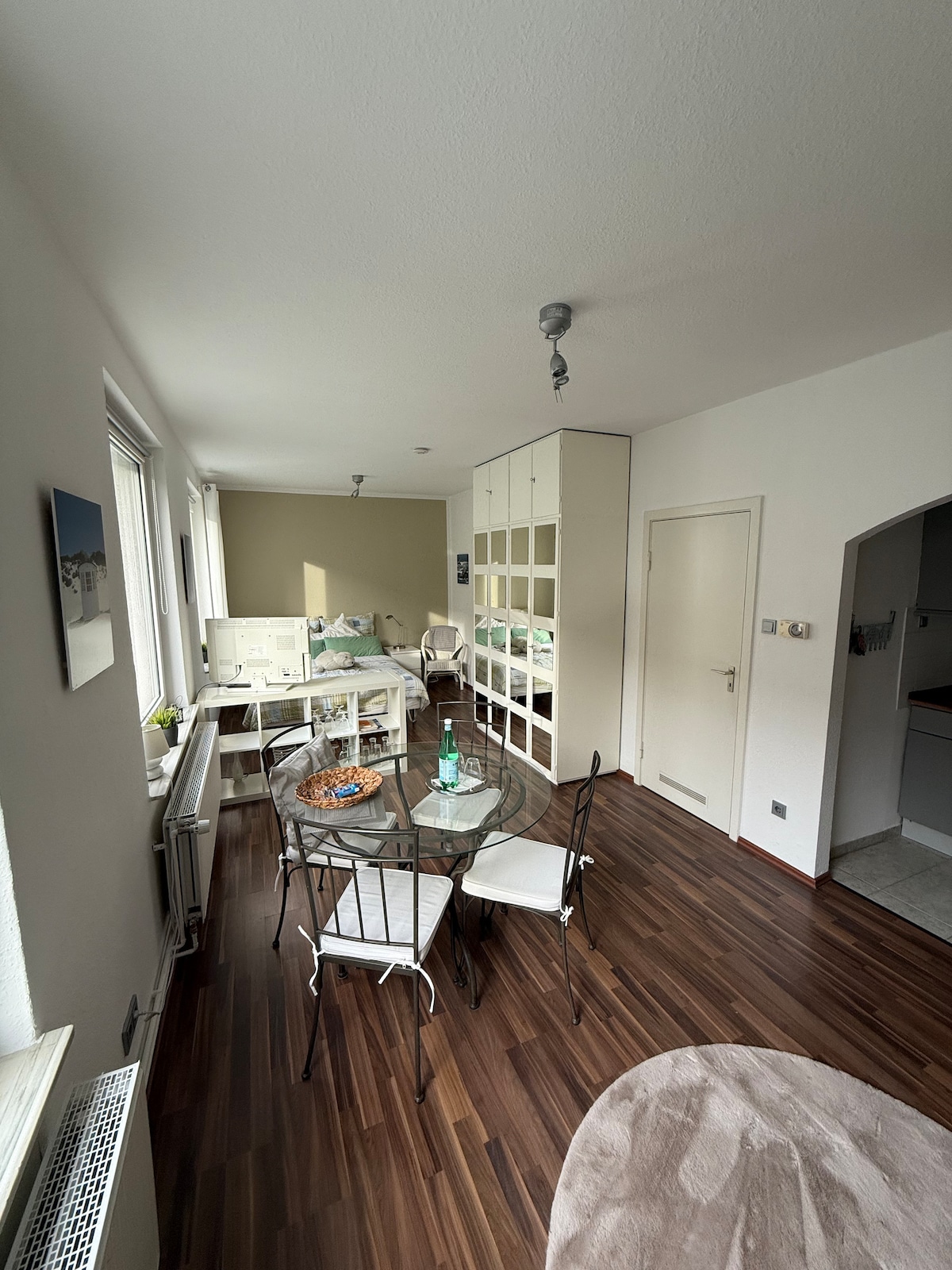 The bright living area features a glass dining table surrounded by four white chairs. A stylish cabinet lines the wall, providing storage. Natural light enters through multiple windows, illuminating the warm wooden floor. A small kitchen area is visible in the background.