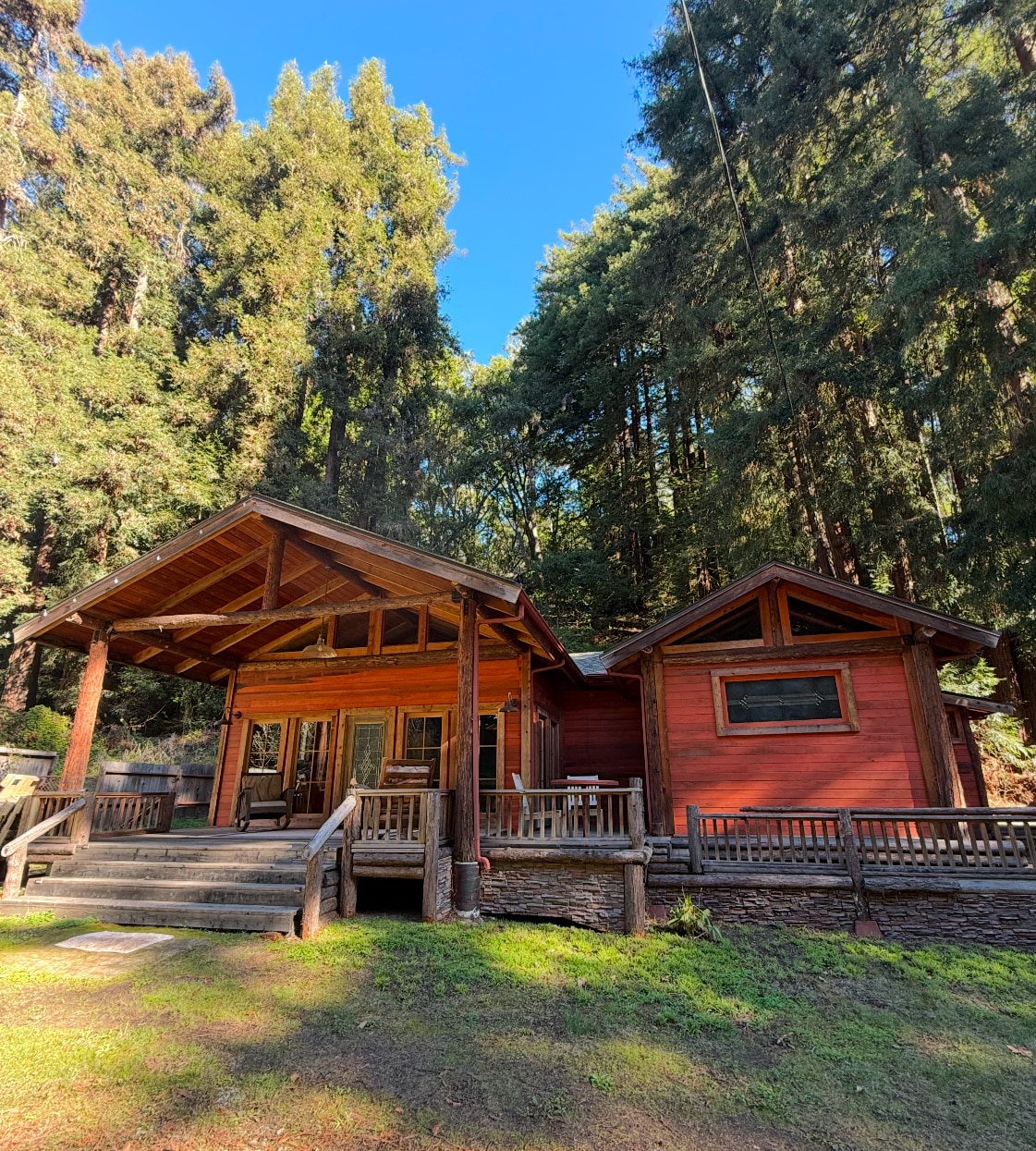 A charming cabin is nestled among tall redwood trees, featuring a wooden deck and two distinct sections. Natural light filters through the surrounding foliage, enhancing the warm wood exterior. The inviting entrance leads to a spacious outdoor area, perfect for relaxation amidst the serene forest setting.