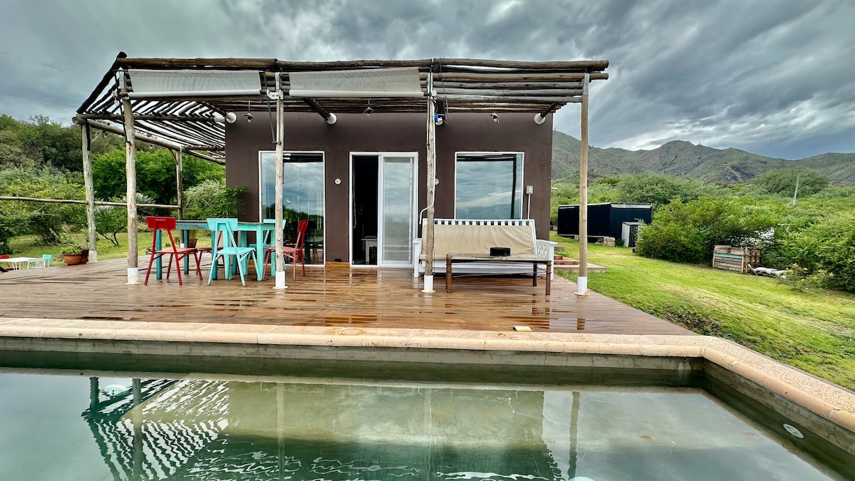 A deck extends from the house, featuring a seating area with a sofa and colorful chairs. Large glass doors provide views of the surrounding nature. The serene pool reflects the cloudy sky, while the green landscape enhances the sense of seclusion.