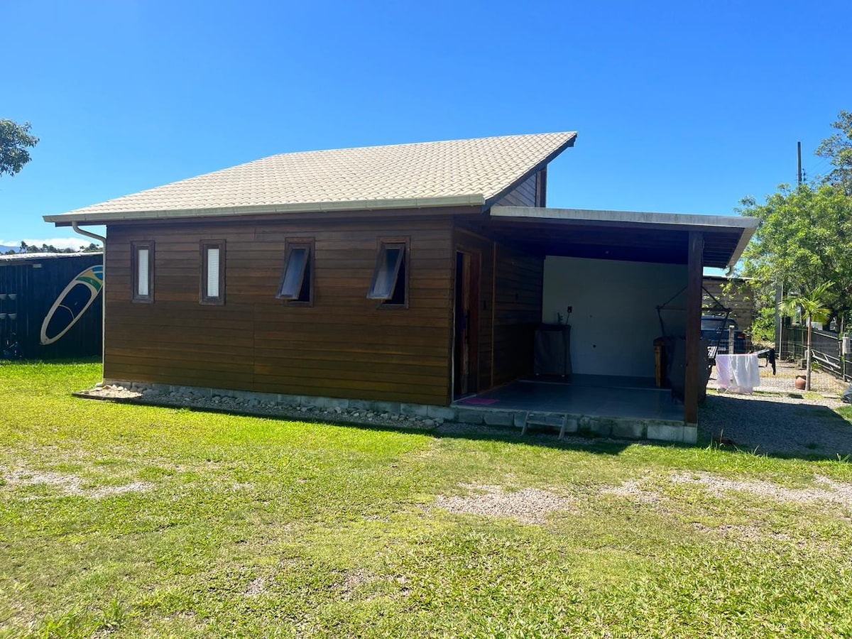 A wooden house is shown with a light-colored roof and multiple small windows. The structure is surrounded by green grass, with a spacious outdoor area visible. The front porch features a concrete surface, providing easy access to the interior.