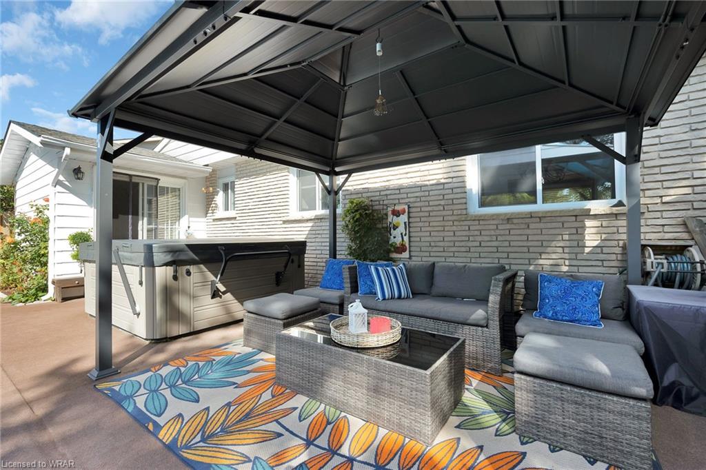 A sheltered outdoor seating area features a sectional sofa adorned with decorative pillows. A coffee table sits in the center on a colorful rug, while a hot tub is positioned nearby. Light filters through the structure, illuminating the relaxed atmosphere.
