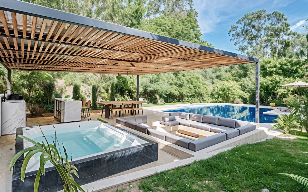 An expansive outdoor area features a covered dining space and a large seating arrangement surrounding a fire pit. A private jacuzzi and swimming pool are visible, framed by lush greenery and sunlight filtering through the trees.