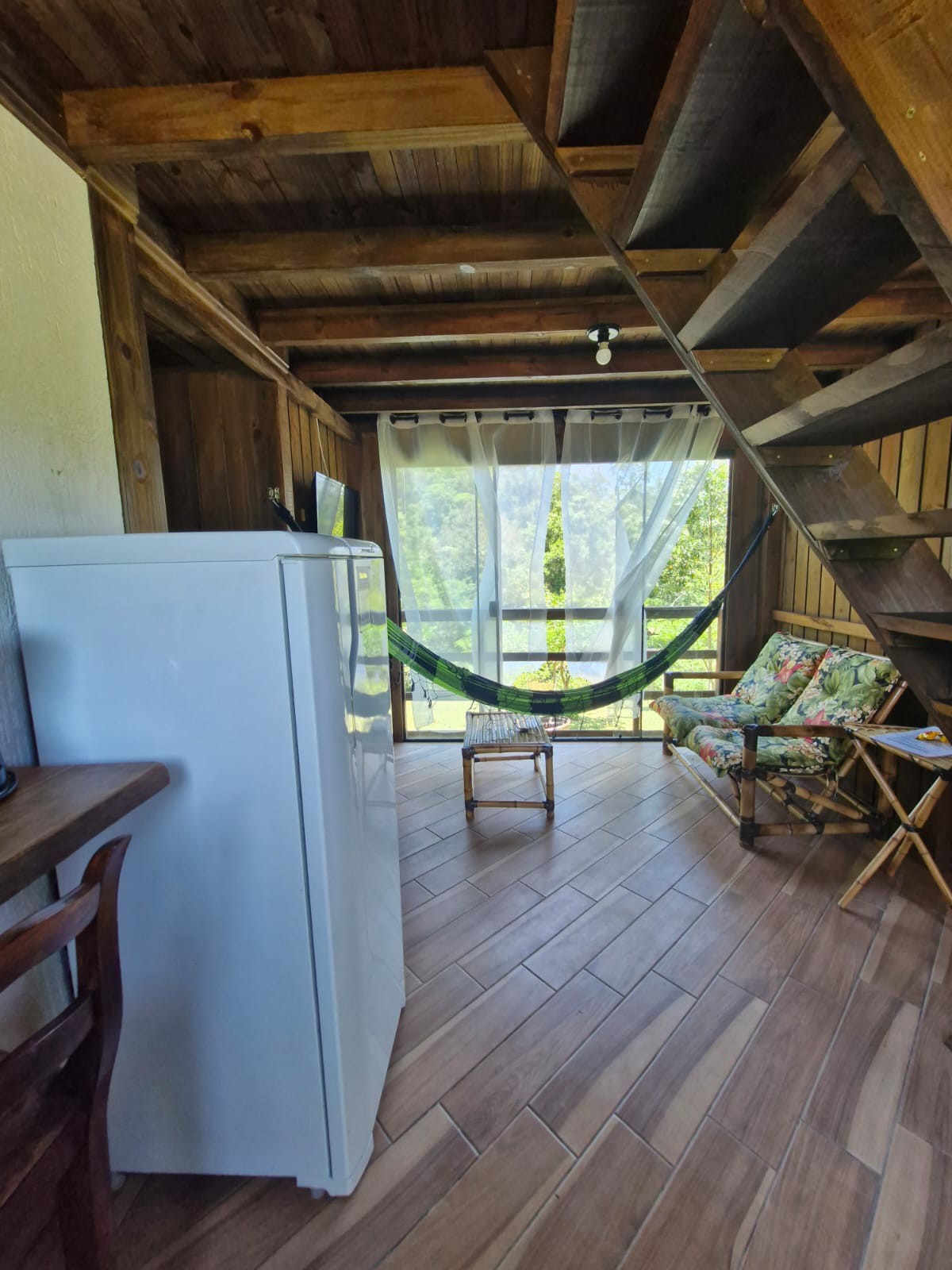 A rustic interior features wooden walls and a tiled floor. A white refrigerator stands beside a wooden table with chairs. A hammock hangs beside a floral-patterned chair, offering a cozy spot to relax. Large windows allow natural light to fill the space while framing views of the surrounding greenery.