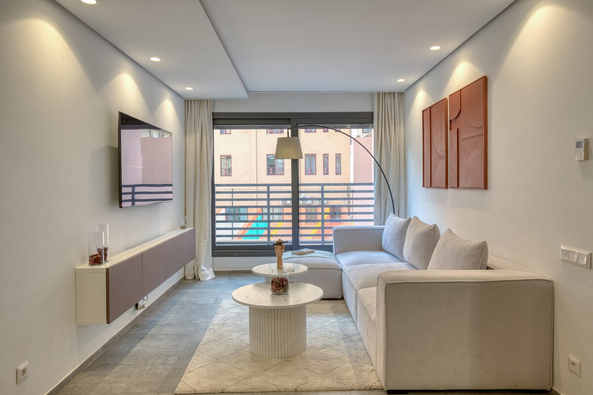 A contemporary living space is highlighted by a light-colored sectional sofa and a round coffee table. A flat-screen television is mounted on the wall, with soft lighting creating a calm ambiance. Large windows allow natural light to enter, offering views of the surrounding area.