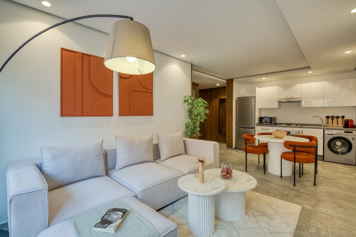 A modern living area is showcased, featuring a light-colored sectional sofa with numerous cushions. A floor lamp stands nearby, casting a soft glow. A round coffee table with decorative items sits in the center, and a dining area with orange chairs is visible in the background.