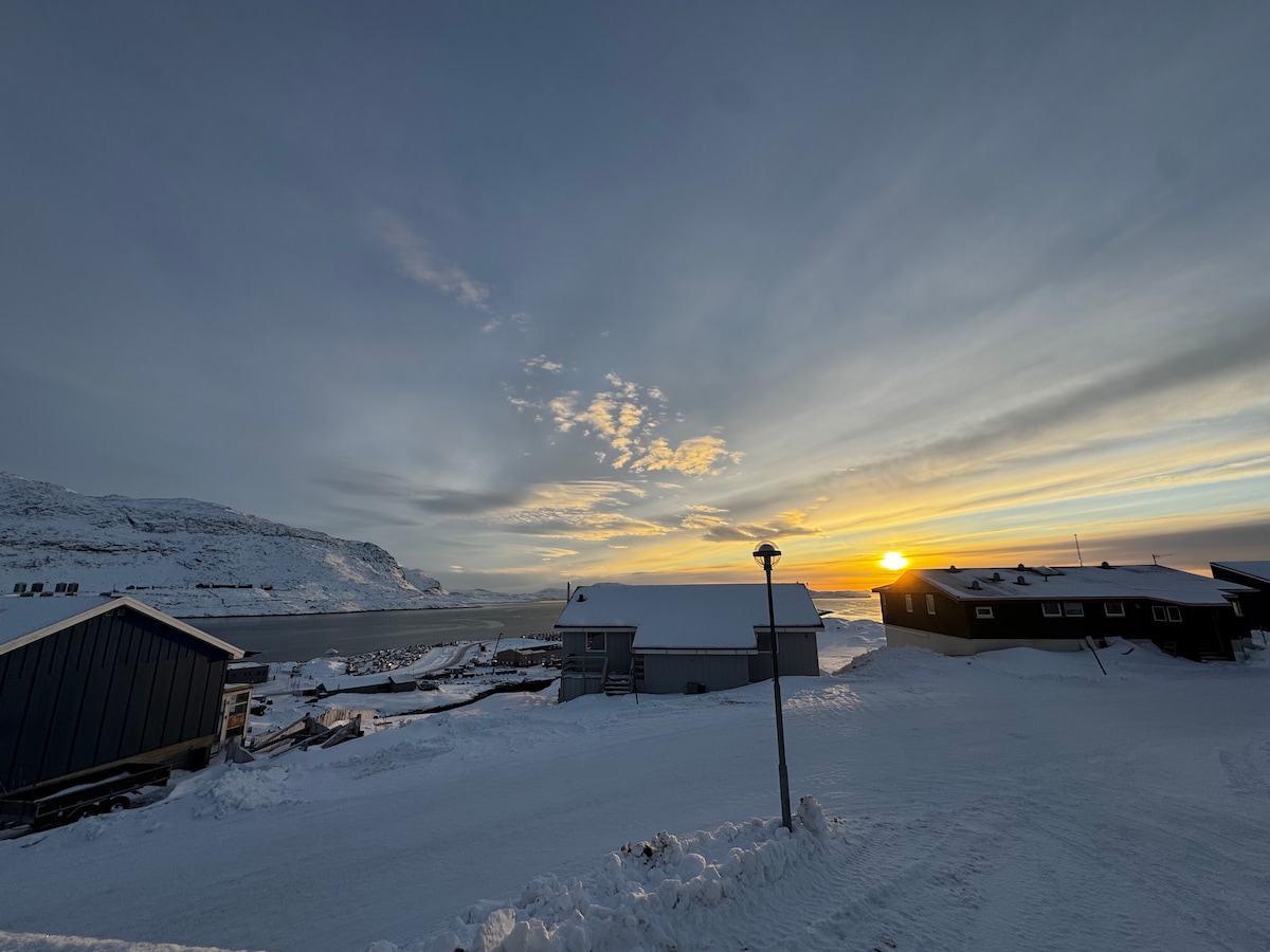 A serene winter landscape showcases a snow-covered expanse leading to calm waters. The sun is setting, casting soft hues across the sky, while rustic buildings are positioned along the shoreline, framed by distant mountains and a gentle sea.