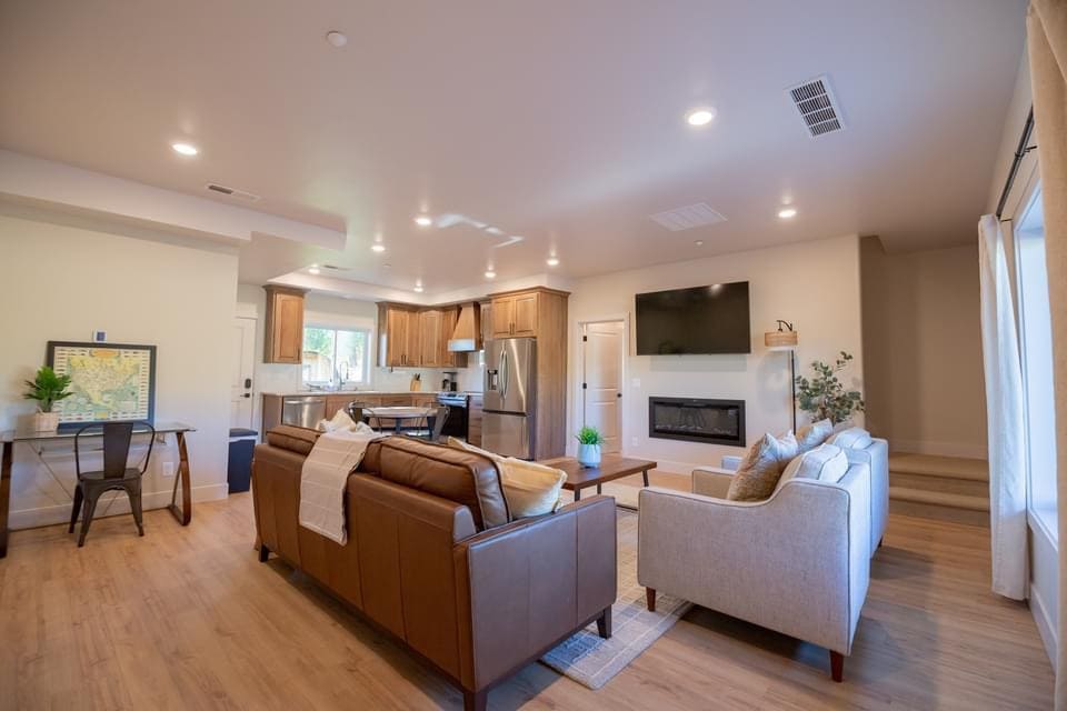 A spacious living area features two comfortable sofas, a coffee table, and a dining table for four. Natural light is welcomed through large windows. The kitchen is visible, showcasing modern appliances and cabinetry. A wall-mounted television is positioned above a contemporary fireplace.