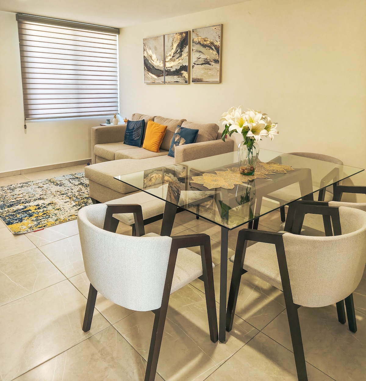 A bright living and dining area is presented, featuring a glass dining table surrounded by four upholstered chairs. A spacious sofa with decorative cushions sits against a wall, complemented by a modern rug and two abstract art pieces above the sofa.