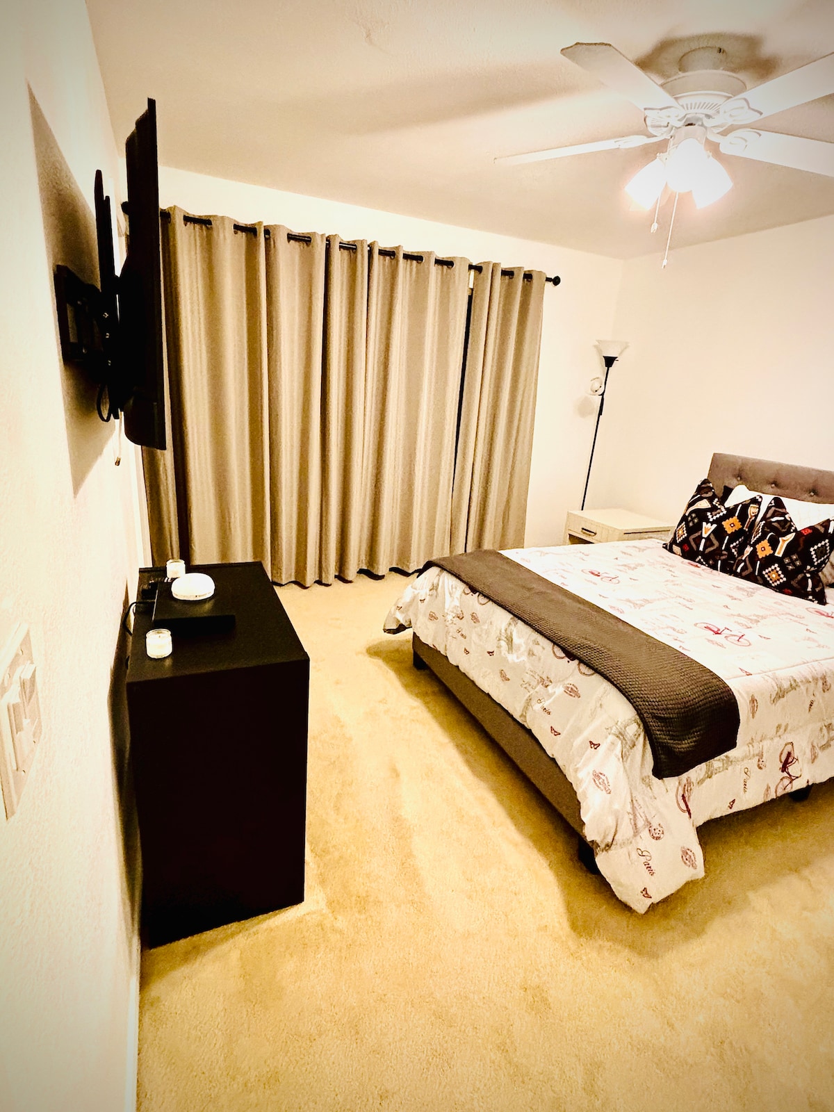 A well-lit bedroom features a spacious bed dressed in patterned bedding. A TV is mounted on the wall opposite the bed. Soft curtains frame a large window, while a lamp and a bedside table are positioned beside the bed, creating a functional layout.
