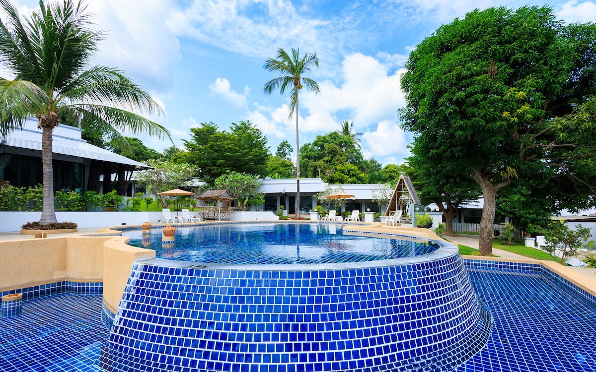 A serene pool area is surrounded by lush greenery and palm trees, featuring shimmering blue tiles. Lounge chairs and umbrellas are positioned on the deck for relaxation. The backdrop includes the resort's elegant buildings, providing a sense of tranquility in the outdoor space.