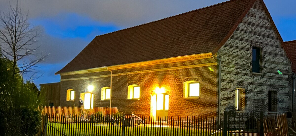 An exterior view of a charming brick and flint barn is presented, showcasing warm lights emanating from several windows. The building features a traditional roofline and is framed by a neatly maintained lawn and fence, creating an inviting atmosphere during twilight hours.