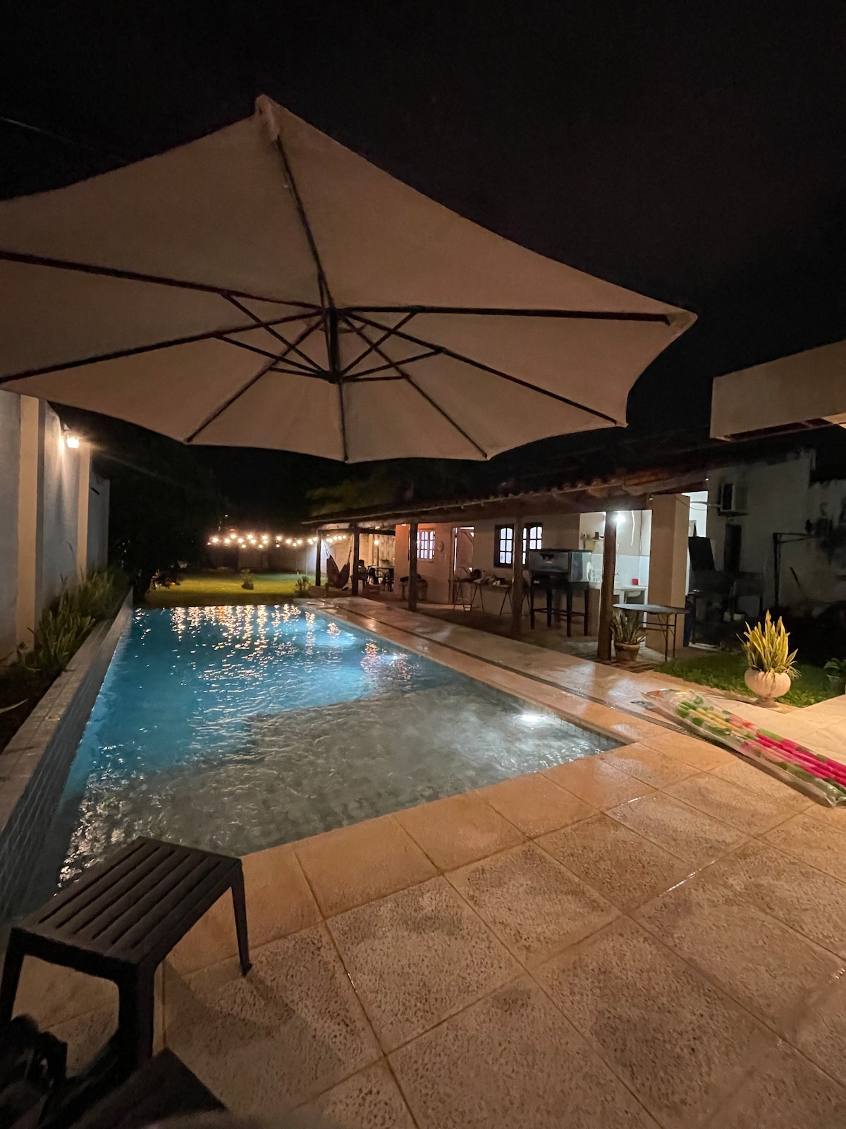The image shows a tranquil pool area illuminated softly at night, featuring a swimming pool with a subtle glow. A large umbrella provides shade over a dark chair. Strings of lights are visible, enhancing the outdoor setting beside the house.