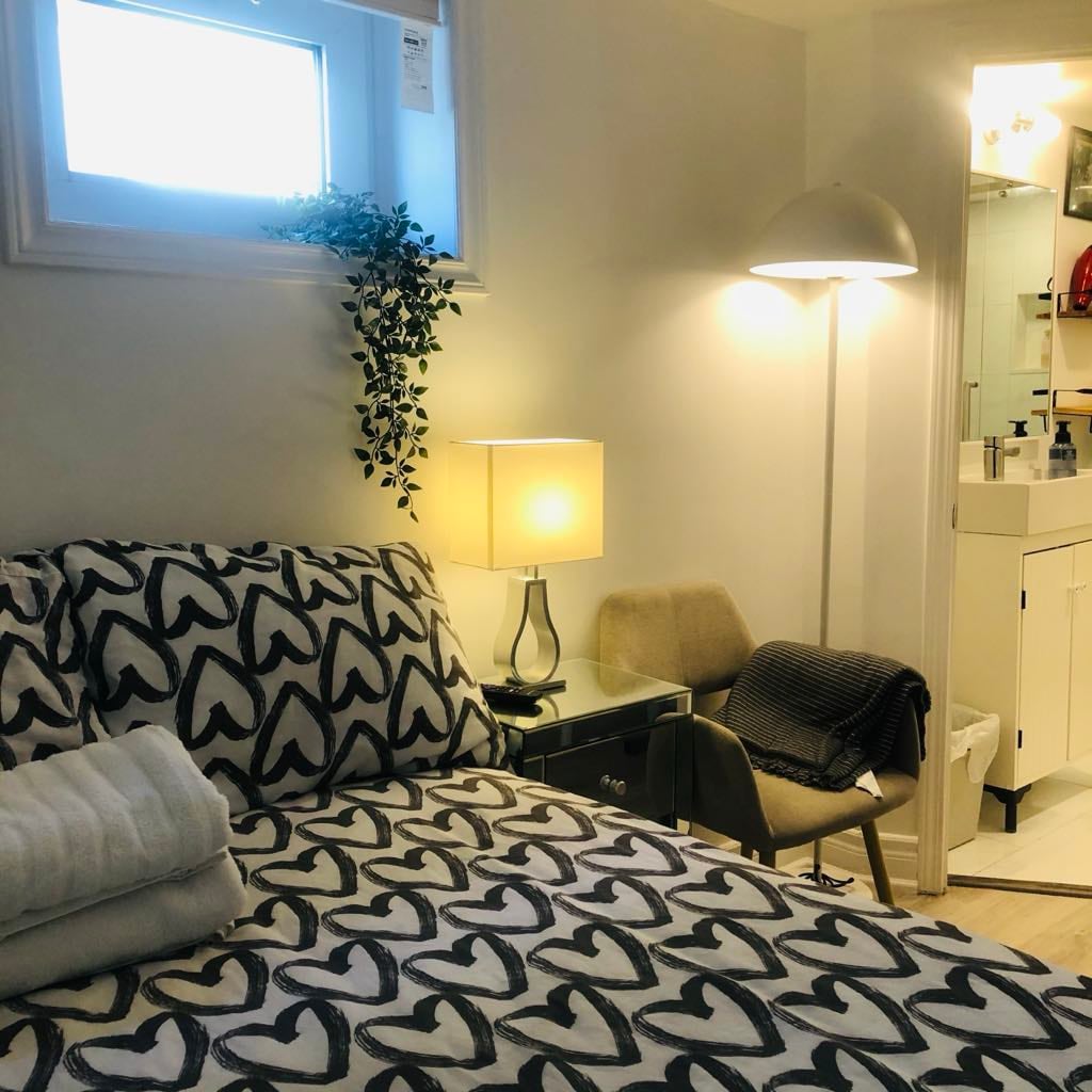 The cozy sleeping area features a double size bed adorned with patterned bedding. A bedside lamp provides soft illumination, and a small chair is positioned nearby. The private bathroom is accessible from the room, with a small window allowing natural light.